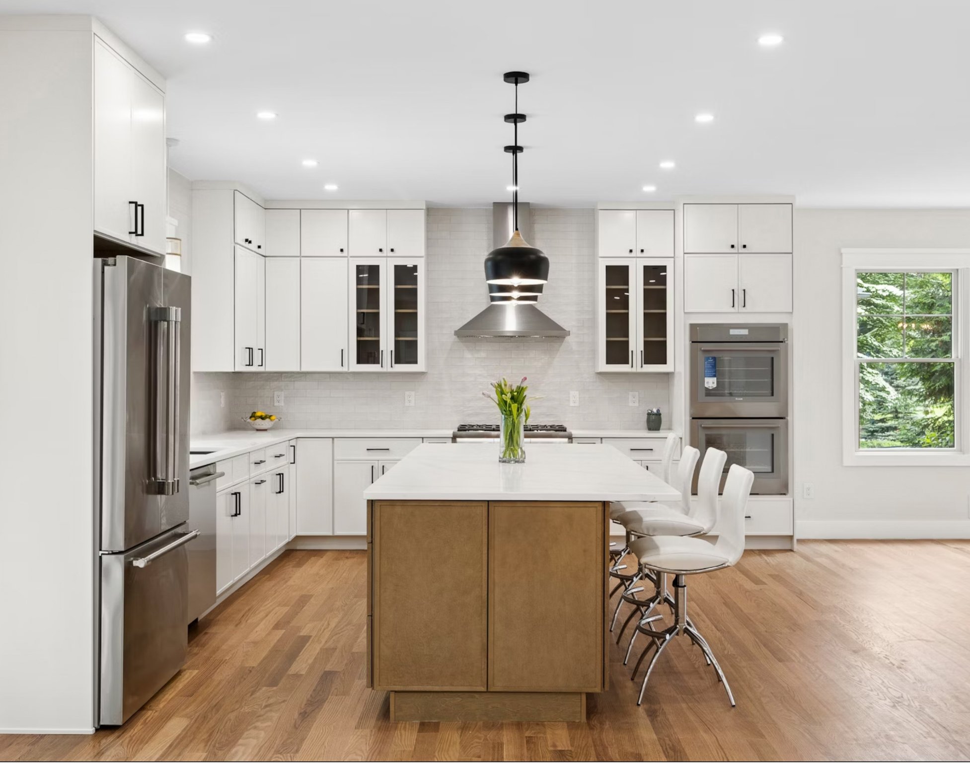 Modern kitchen with white cabinets, a central island with a wooden base, and white bar stools. Contains stainless steel appliances, a stainless steel range hood, and a marble countertop. Green plants are styled in a glass vase on the island. Hardwood