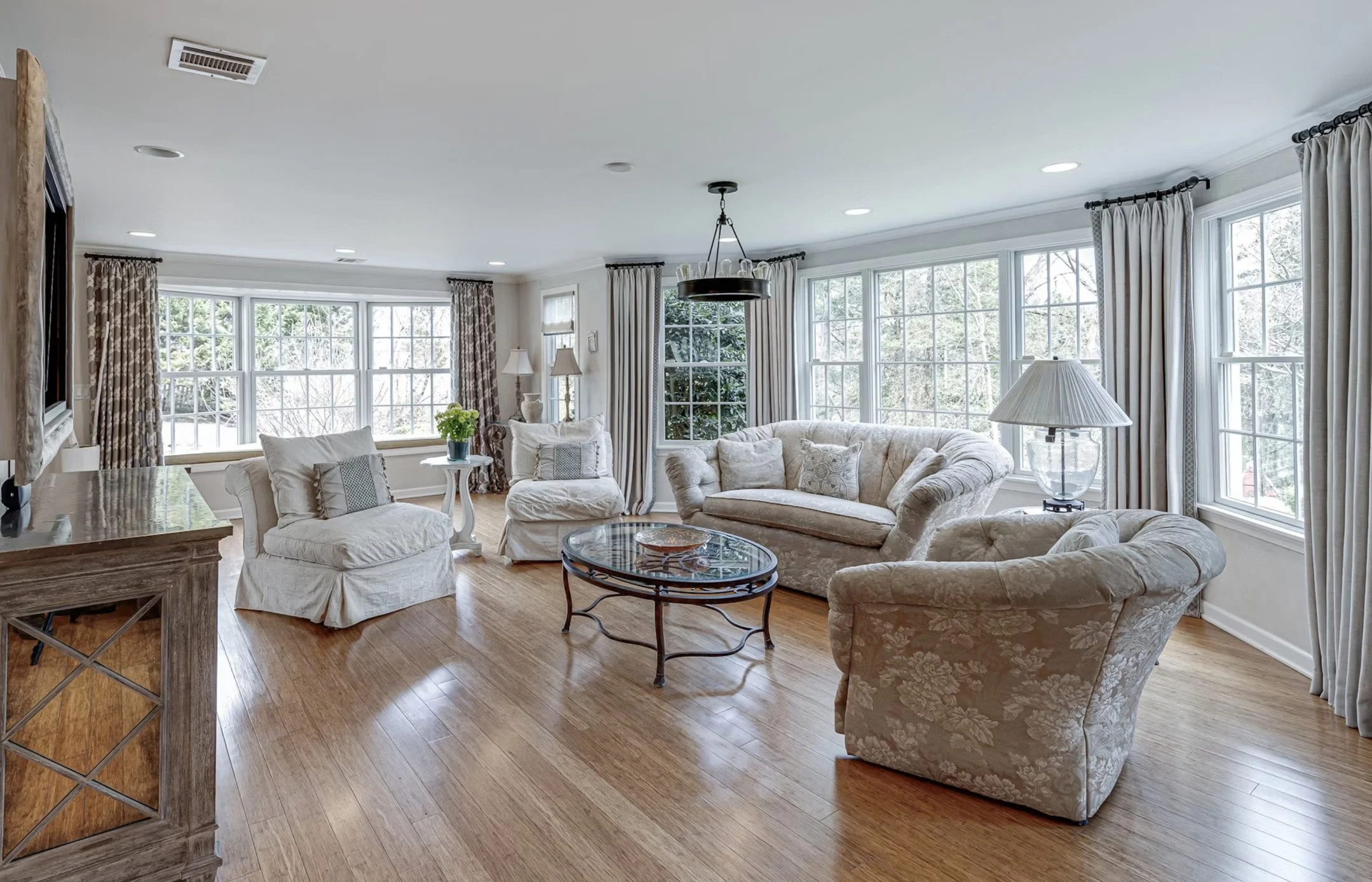 Bright living room with large windows, cream upholstered sofas and armchairs, a wooden coffee table, hardwood floors, and curtains.