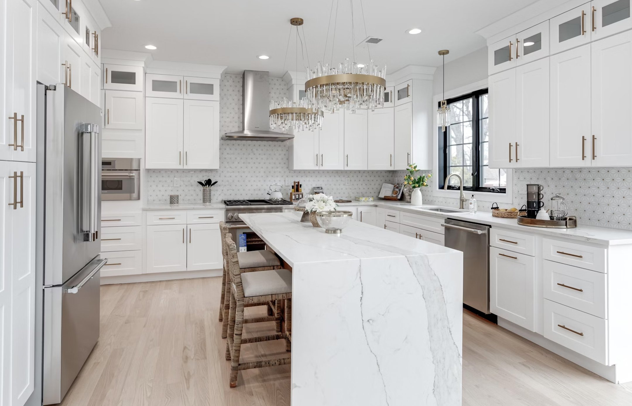 Modern white kitchen with marble island, stainless steel refrigerator, and gold cabinet handles. Lighting fixtures with glass and gold accents hang from the ceiling. Window above the sink shows outdoor trees.