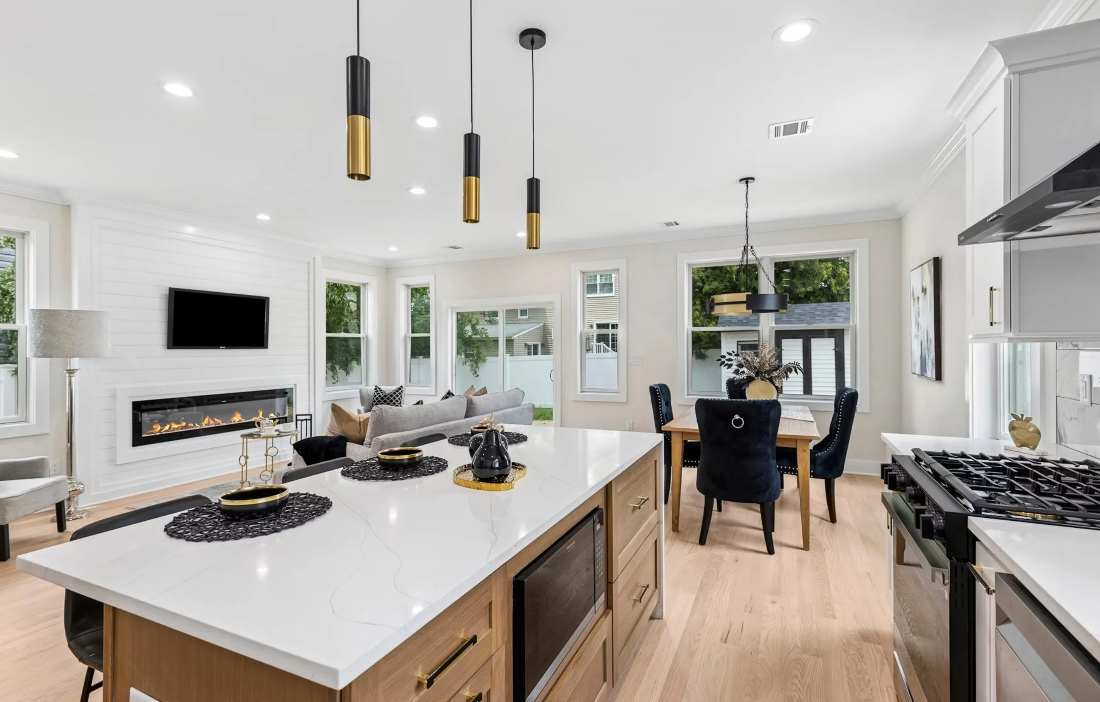 Open-concept kitchen and living room with white walls, hardwood floors, large windows, modern fireplace, gray sofa, dining table with black upholstered chairs, and pendant lighting.