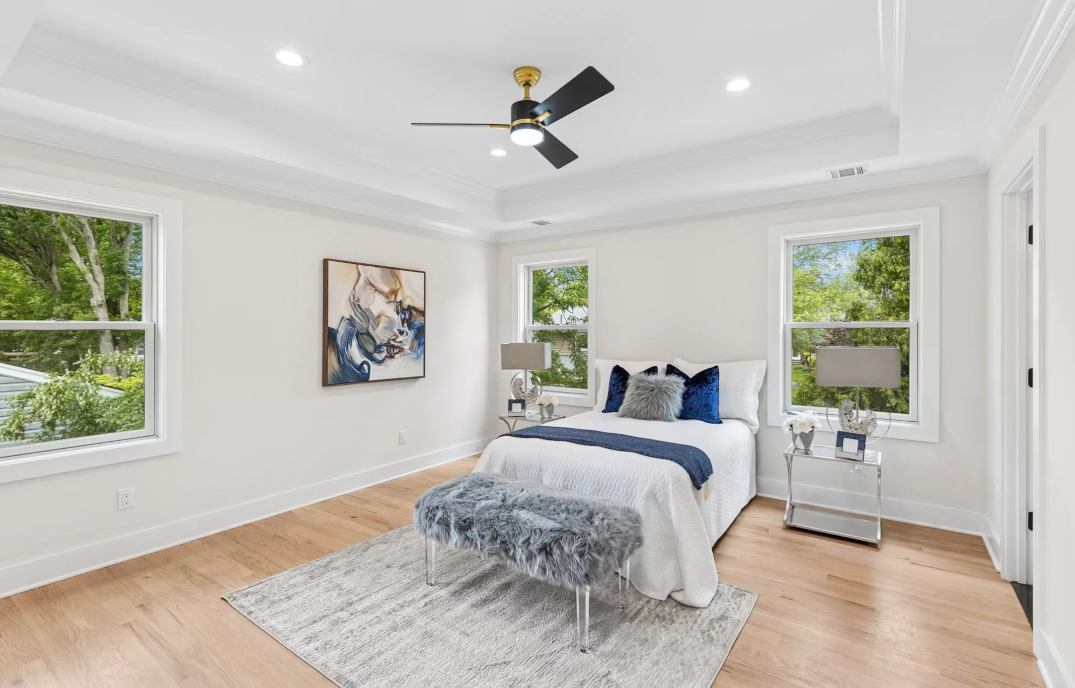 Bright bedroom with white walls, wooden floors, a bed with white linens and blue and gray pillows, two windows showing green trees outside, a modern ceiling fan, and two nightstands with lamps.