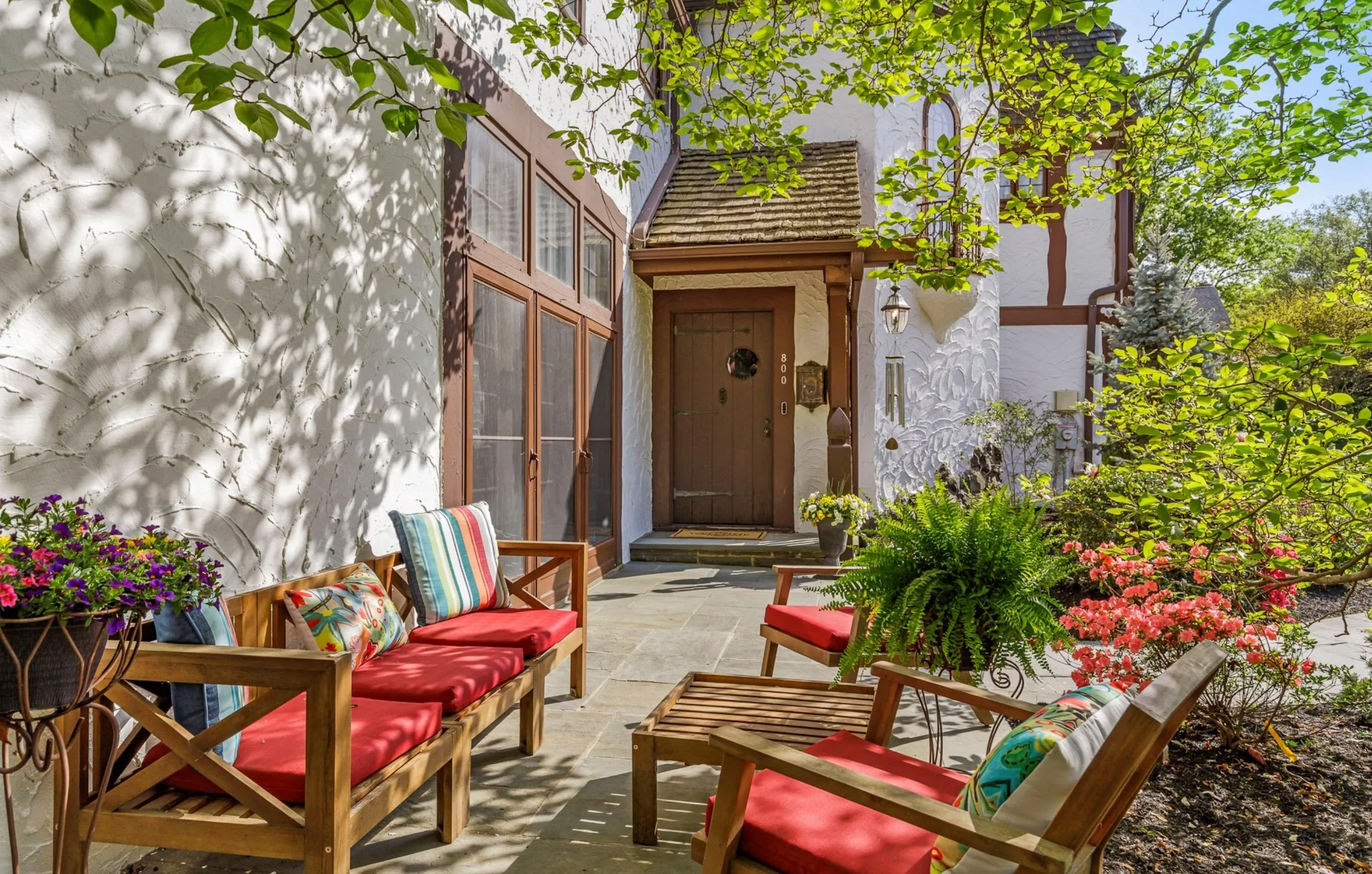 A cozy outdoor patio with wooden furniture and red cushions, flowering plants, and the entrance to a house with a wooden door and windows, surrounded by greenery.