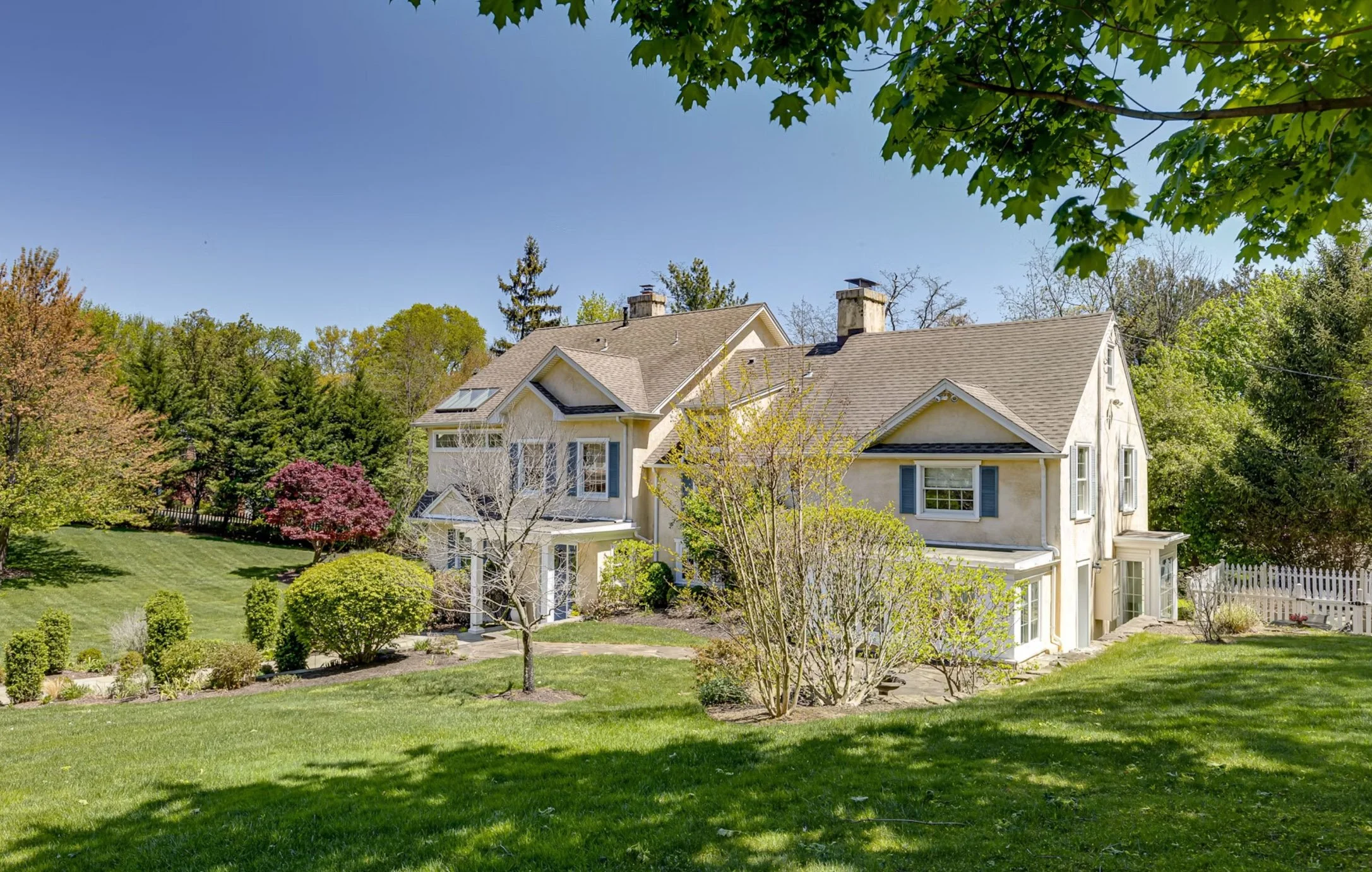 A large, two-story house with a beige exterior surrounded by green trees and a well-manicured lawn, with some trees in bloom and a white picket fence on the right side.