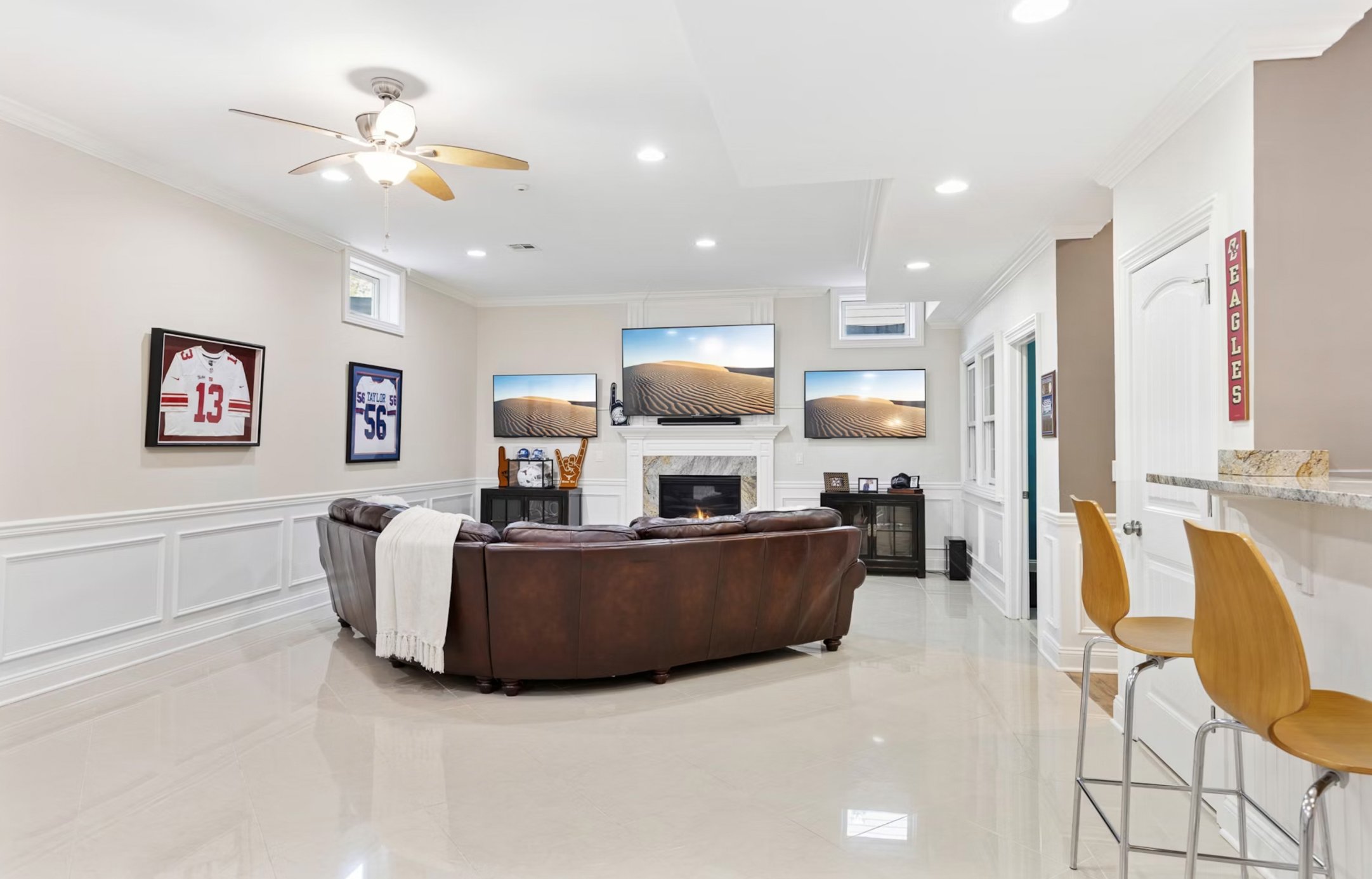 Spacious living room with a leather sectional sofa, three wall-mounted TVs above a fireplace, sports memorabilia on the wall, and bar stools at a breakfast bar.