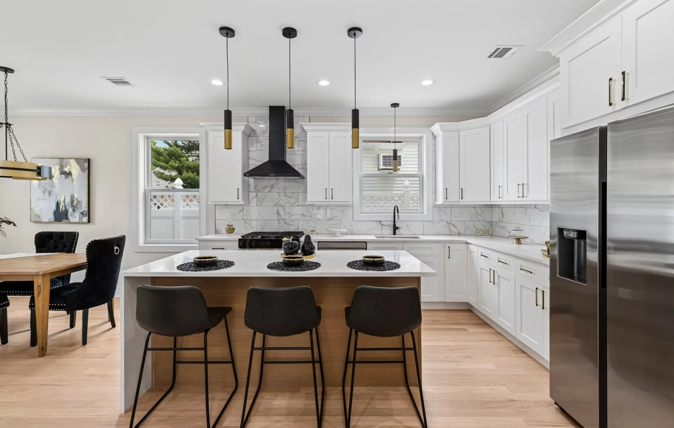 Modern kitchen with white cabinets, marble backsplash, black range hood, and island with black barstools, pendant lights, wooden dining table with black chairs, and stainless steel refrigerator.