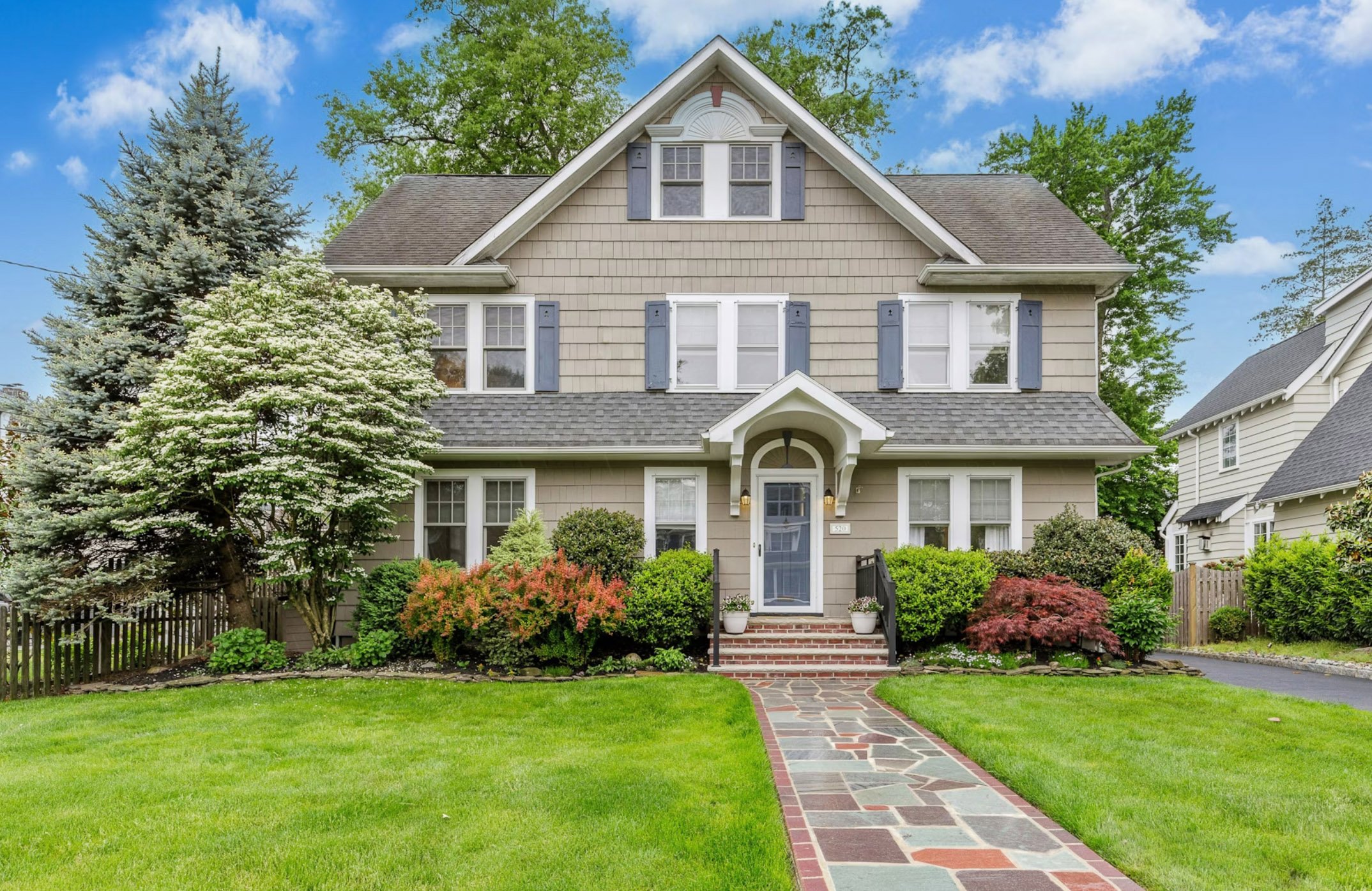 A two-story house with beige exterior, gray roof, and white trim, featuring multiple windows with blue shutters, a small front porch with steps, and a manicured lawn with various shrubs and trees under a blue sky with scattered clouds.