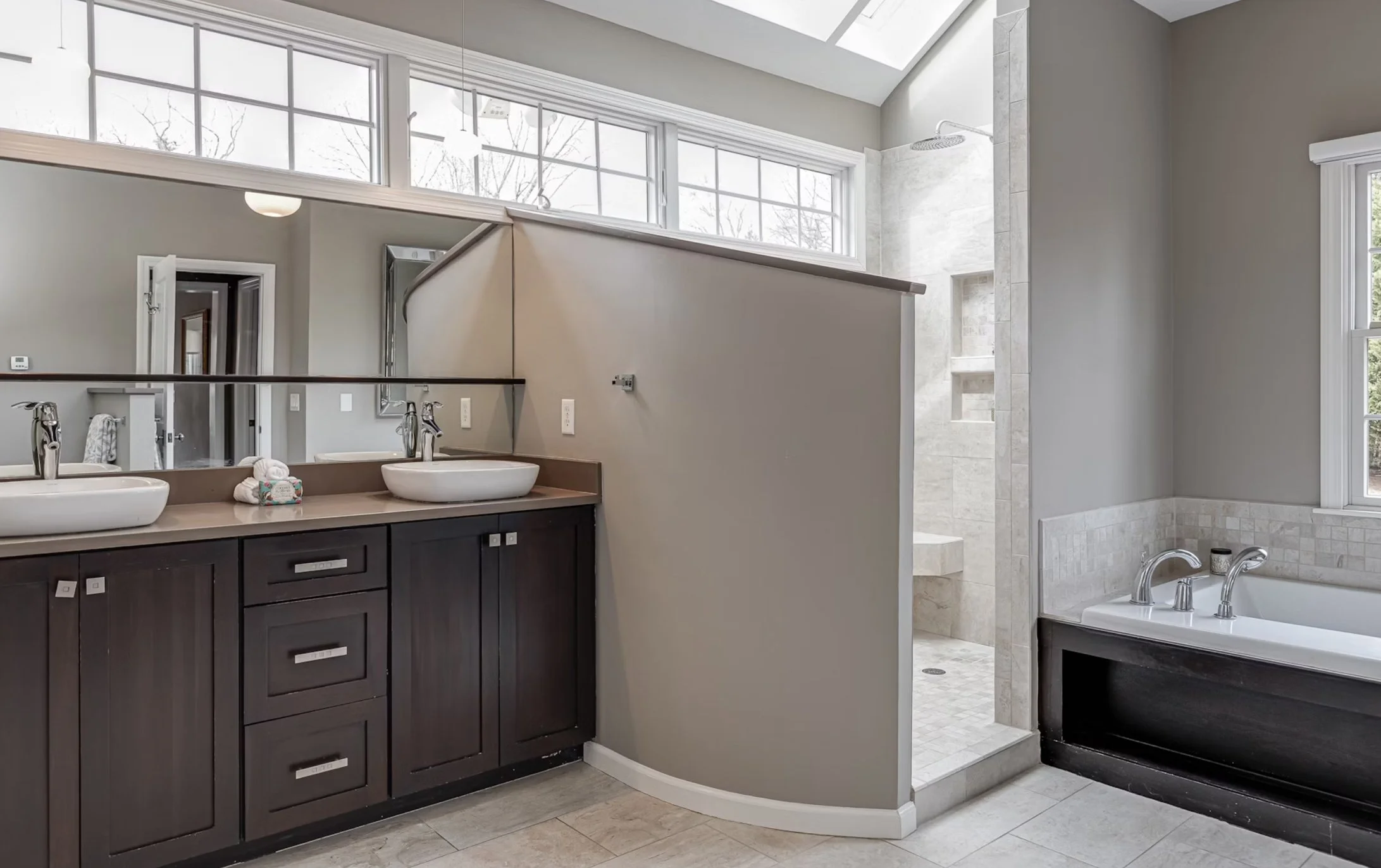 Modern bathroom with a double vanity with vessel sinks, a large mirror, a walk-in shower, and a bathtub with black base trim. Large windows and a skylight provide natural light.