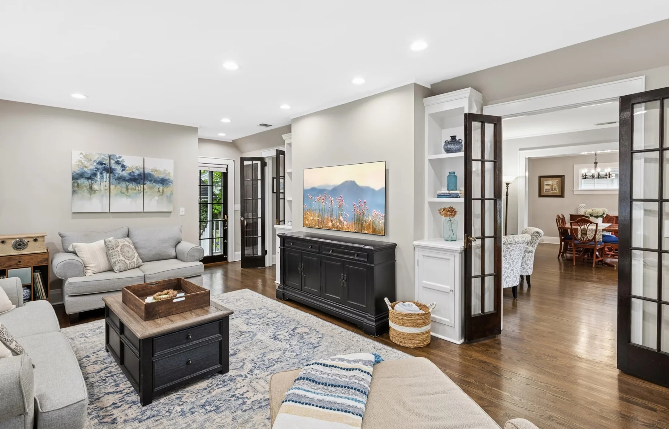 Living room with beige and gray furniture, hardwood floors, and a sliding glass door leading outside. There are wall art pieces, a TV on a black console, and a mix of decorative items.