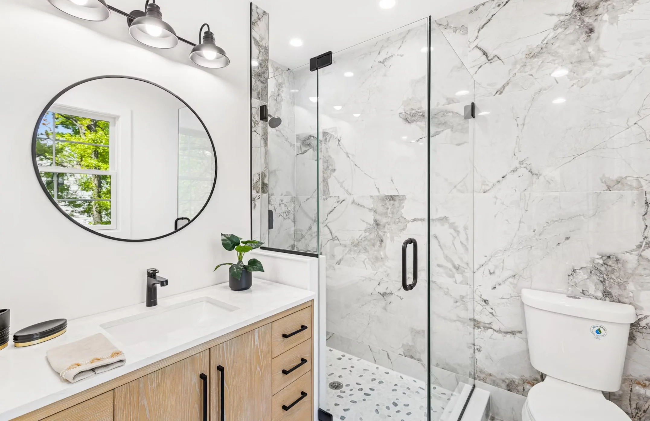 Modern bathroom with white and gray marble walls, a glass shower enclosure, a wooden vanity with a white countertop, a black faucet, a round mirror, a small plant, and a white toilet.