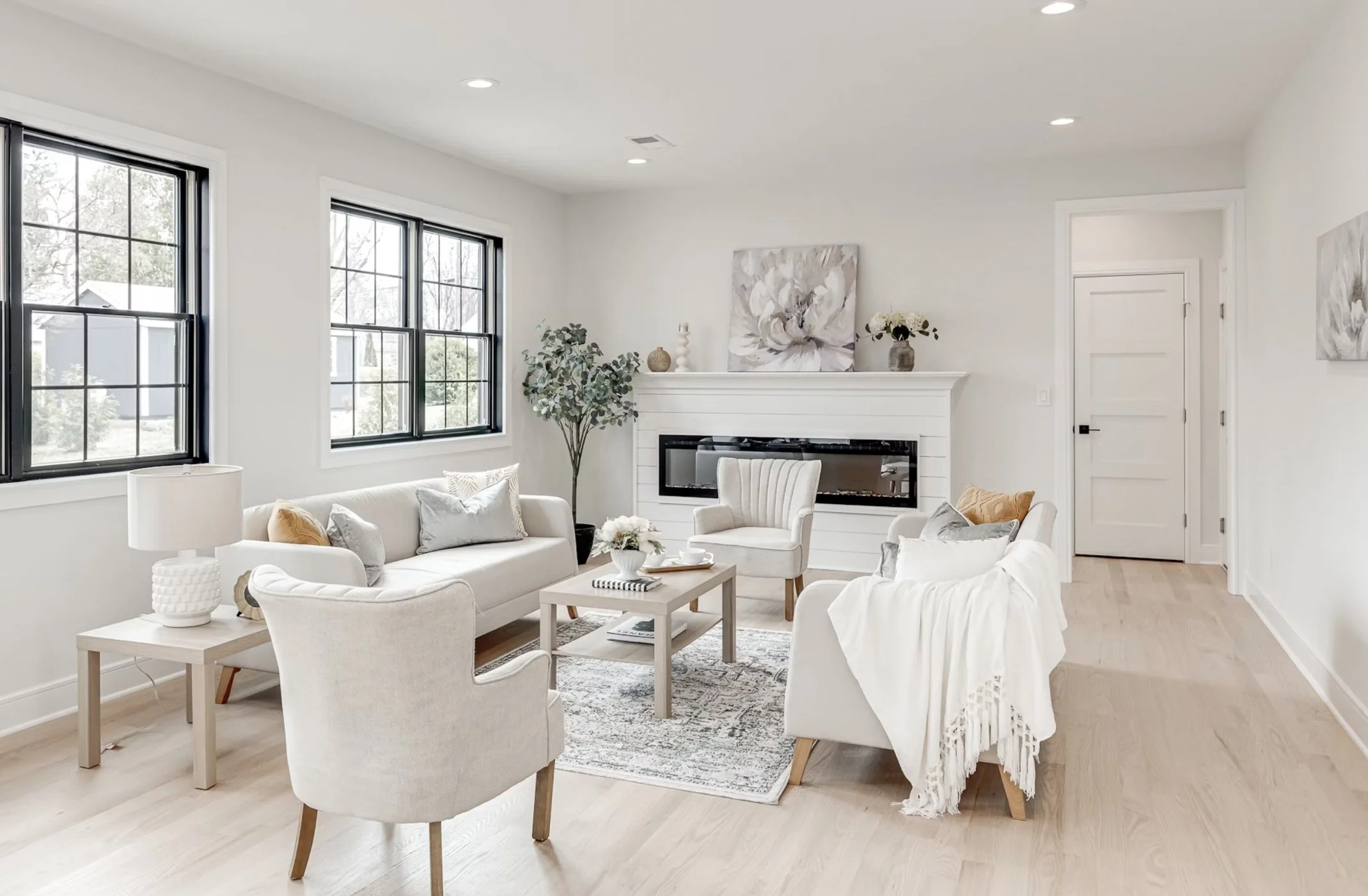 Bright living room with white walls, light wood flooring, two large windows, a white sofa with pillows, two armchairs, a coffee table with flowers, a fireplace, and minimal decorations.