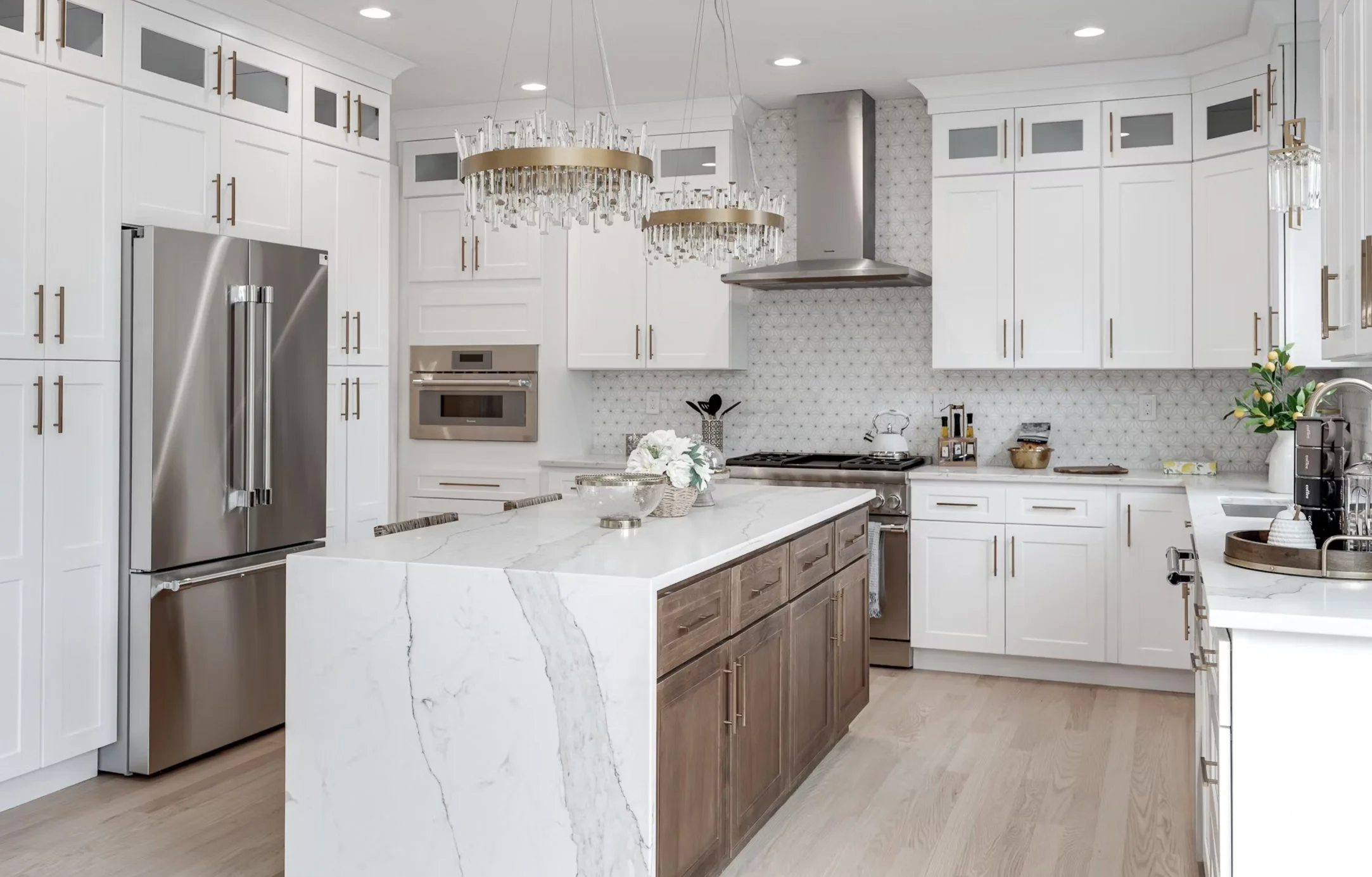 Modern kitchen with white cabinets, stainless steel refrigerator, marble island and wood lower cabinets, gold hardware, pendant chandeliers, patterned backsplash, and various kitchen accessories and plants.