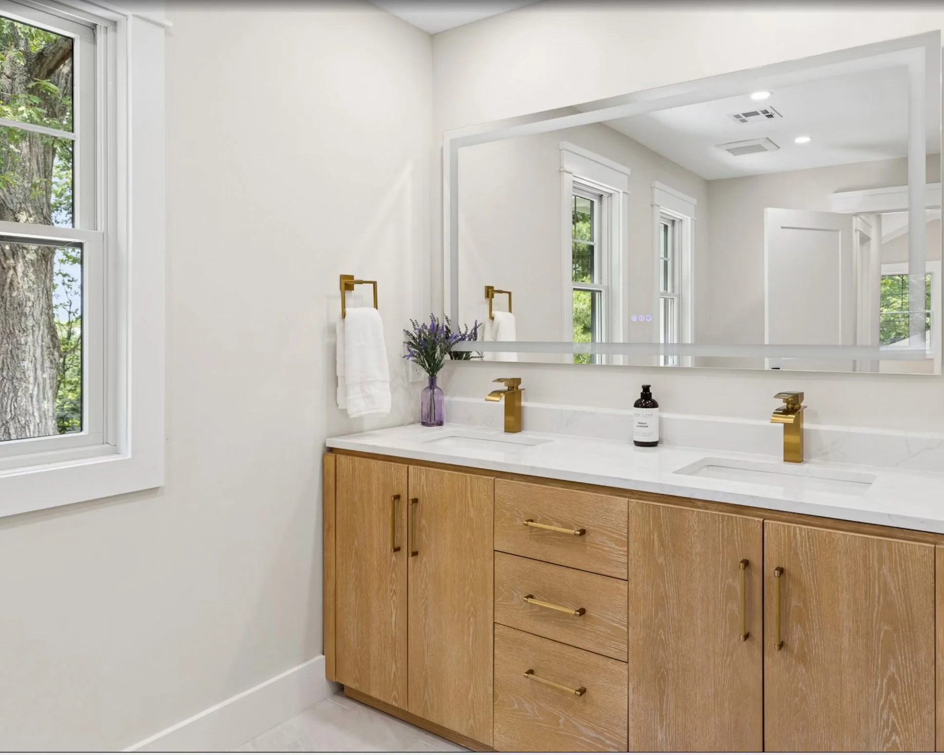 Modern bathroom with wooden vanity, white marble countertop, two golden faucets, large mirror, purple vase with flowers, white towels, window with trees outside, and white walls.