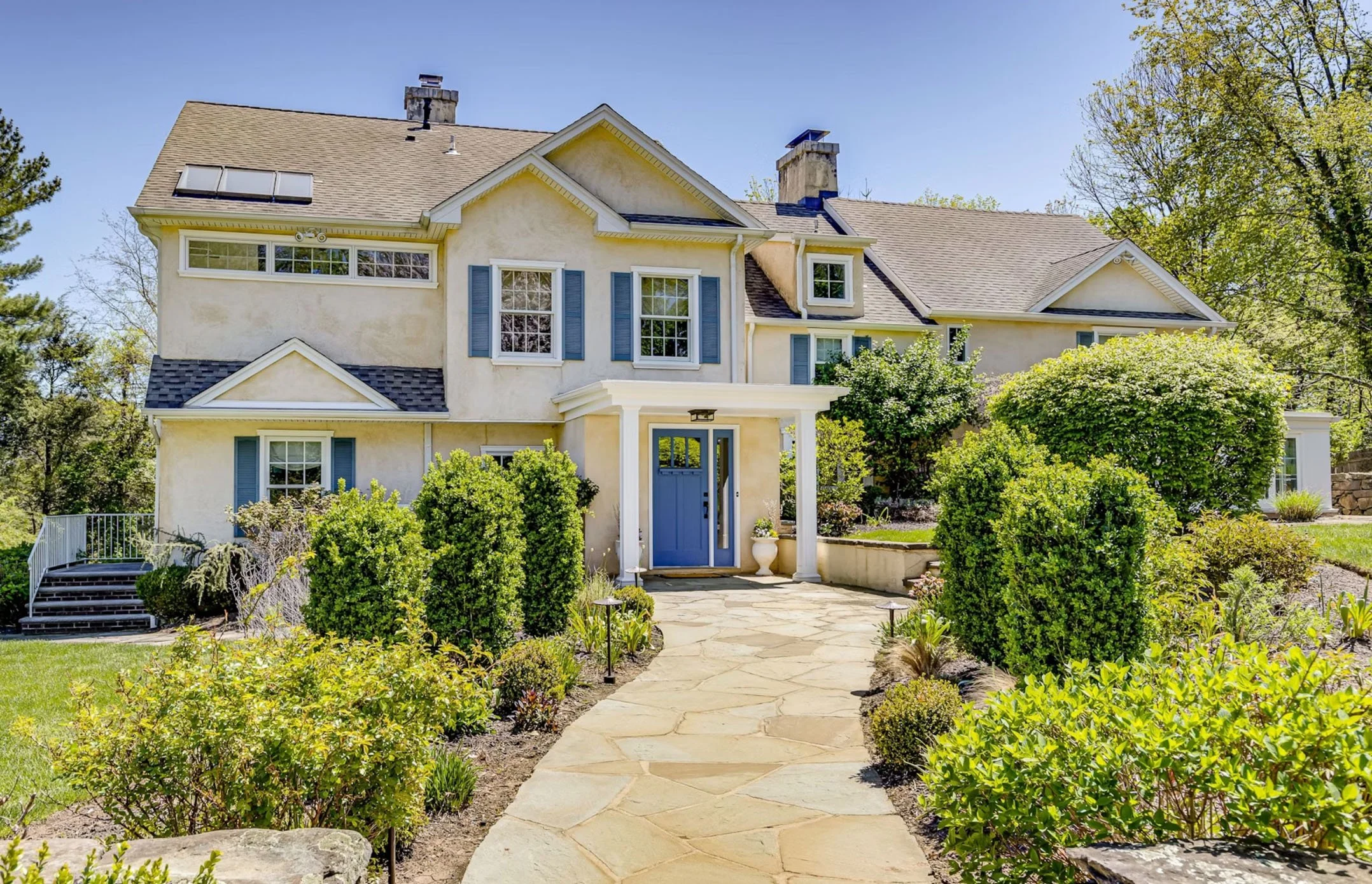 A large, multi-story house with a yellow exterior and blue shutters, surrounded by a landscaped garden and a stone pathway leading to the front door.
