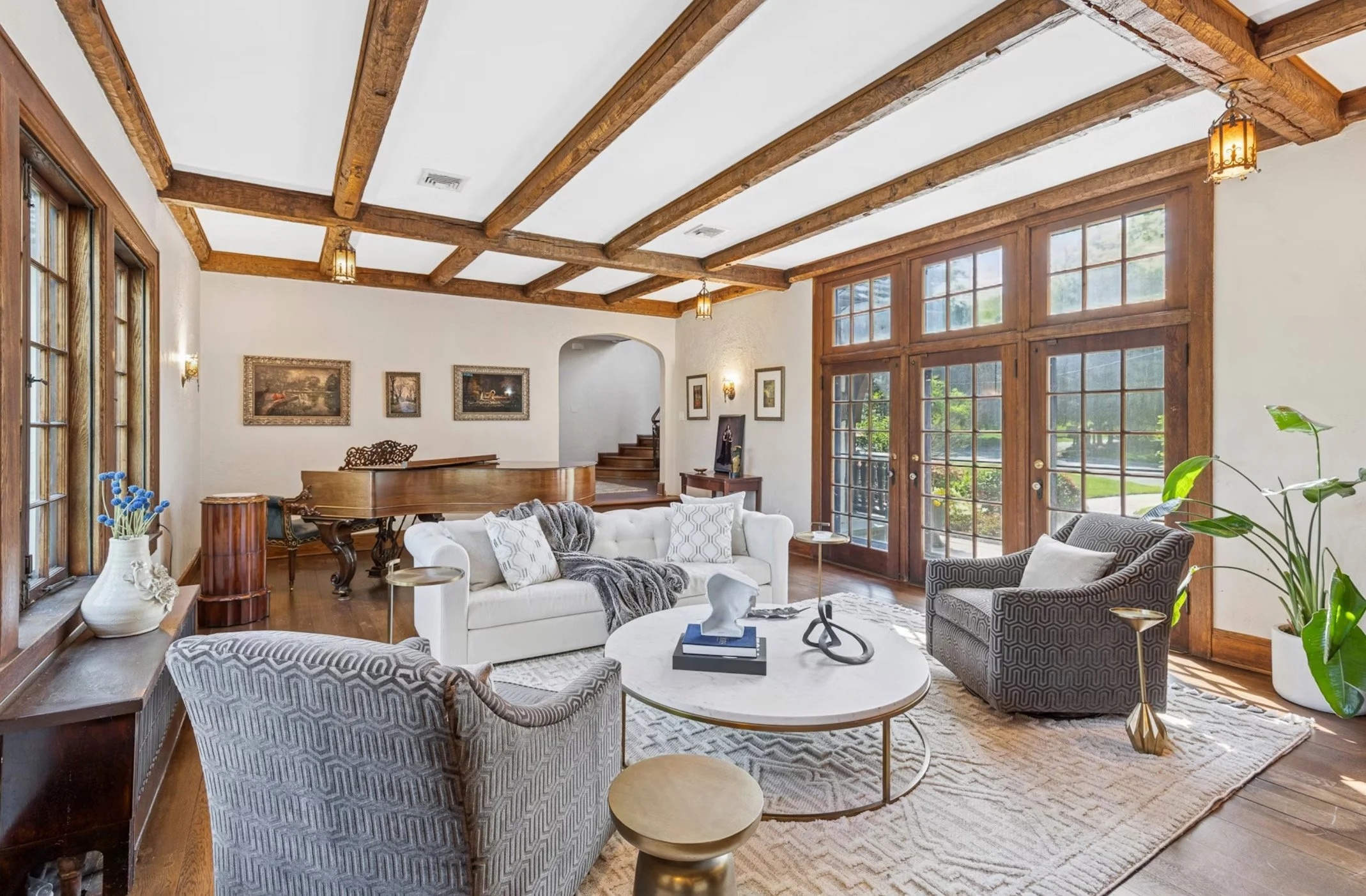 Living room with wooden ceiling beams, large windows, white sofa, patterned armchairs, coffee table with sculptures, potted plants, and artwork on the walls.