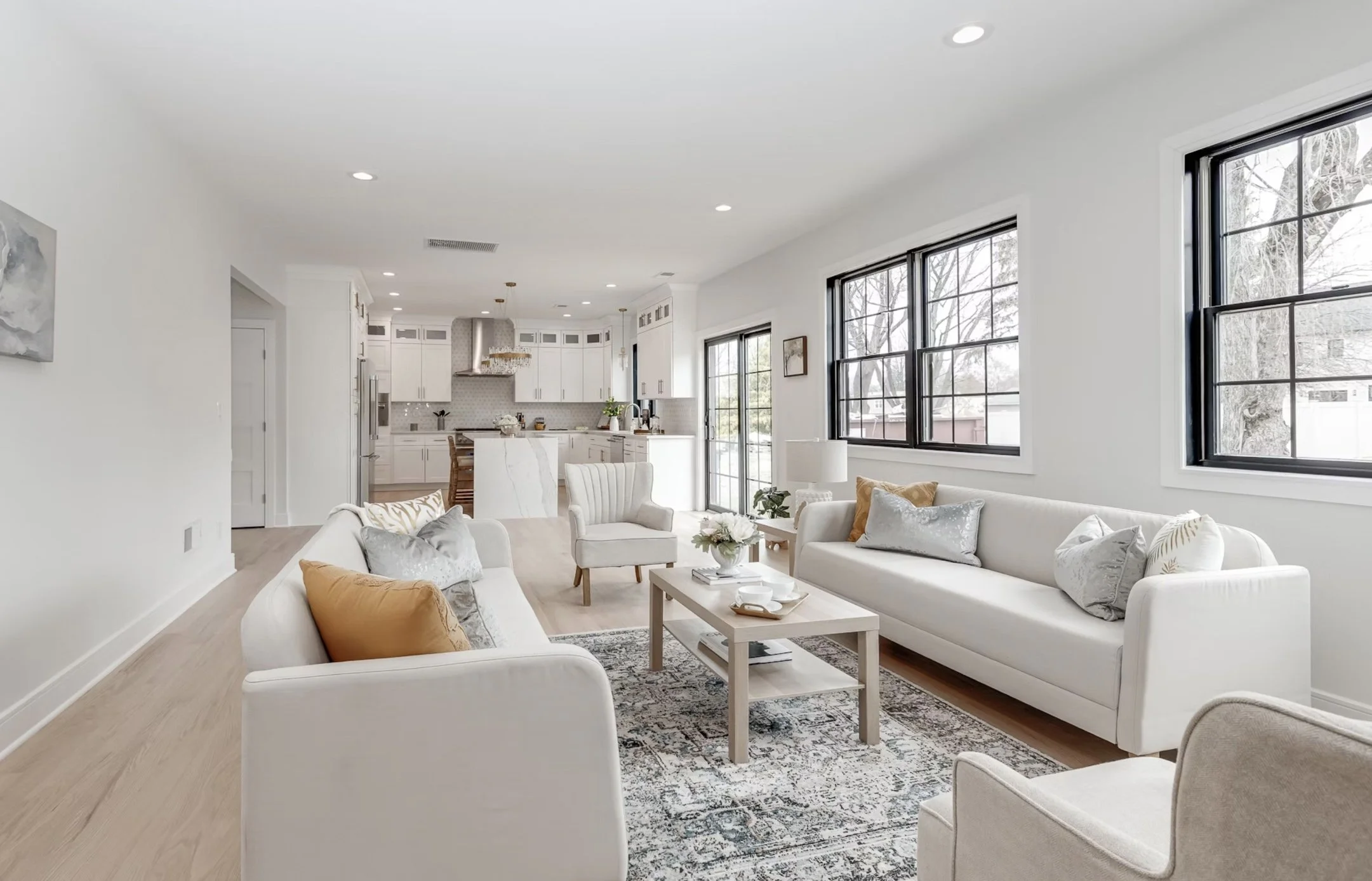 Bright, modern living room with white sofas, armchair, decorative pillows, coffee table, and a patterned rug, with large black-framed windows and a view into the kitchen.