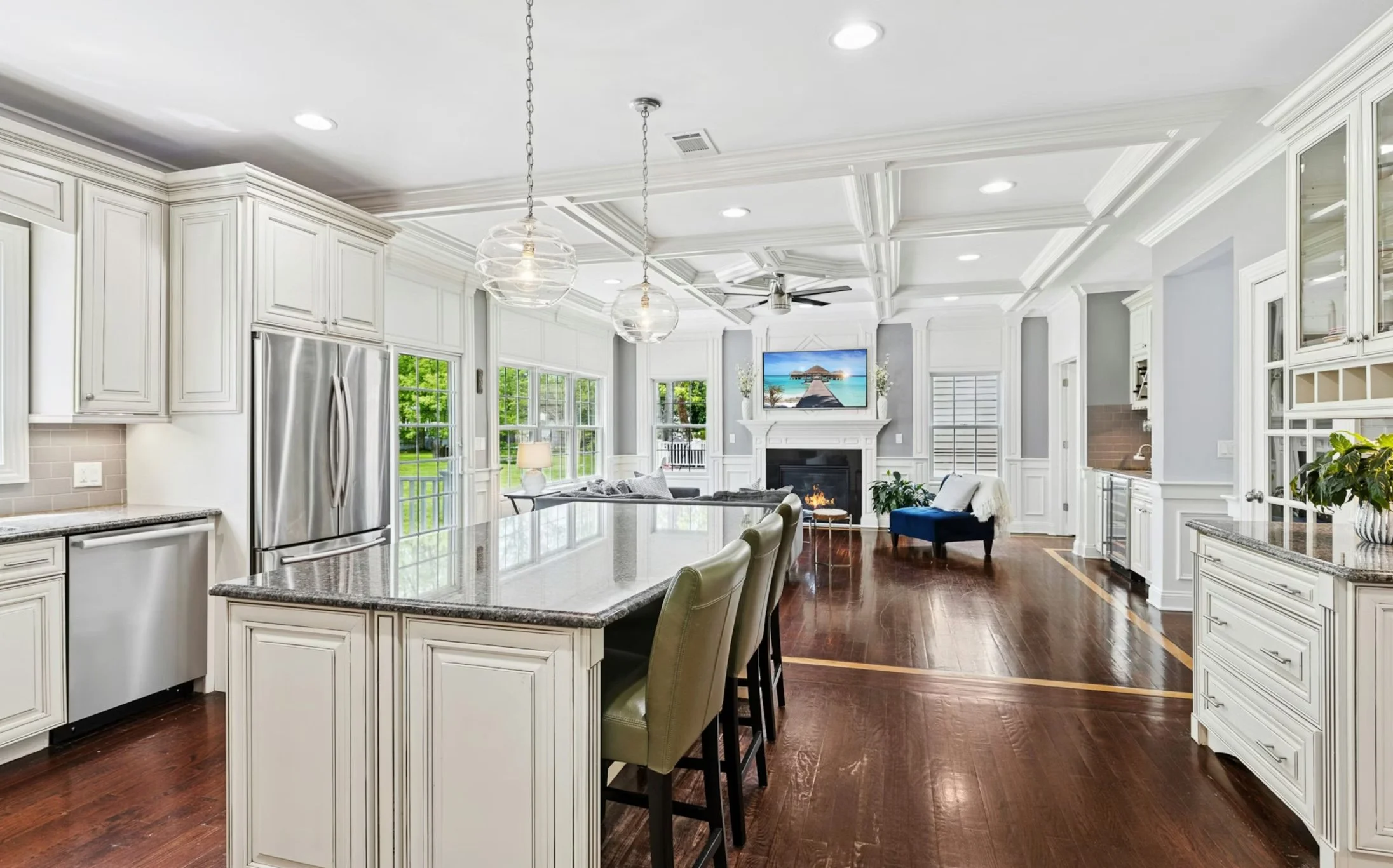Spacious open-concept kitchen and living room with white cabinetry, stainless steel appliances, hardwood floors, large island with seating, and a cozy fireplace with mounted TV, large windows, and classic architectural details.