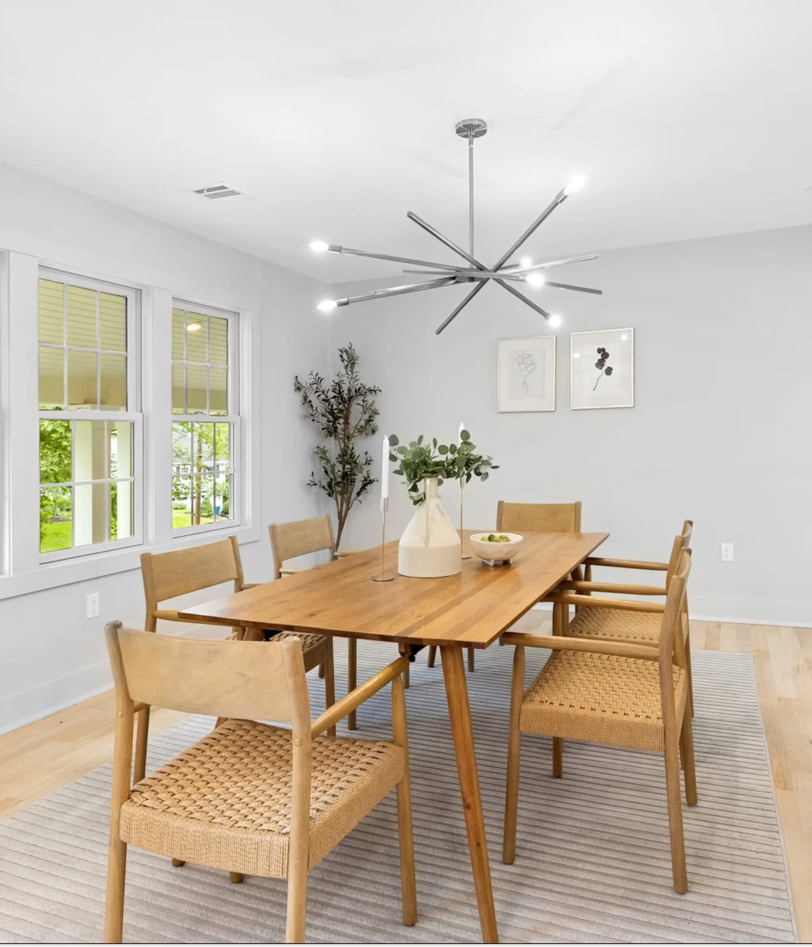 Modern dining room with wooden table, six chairs, white walls, large window, black and white wall art, greenery, and a contemporary chandelier.