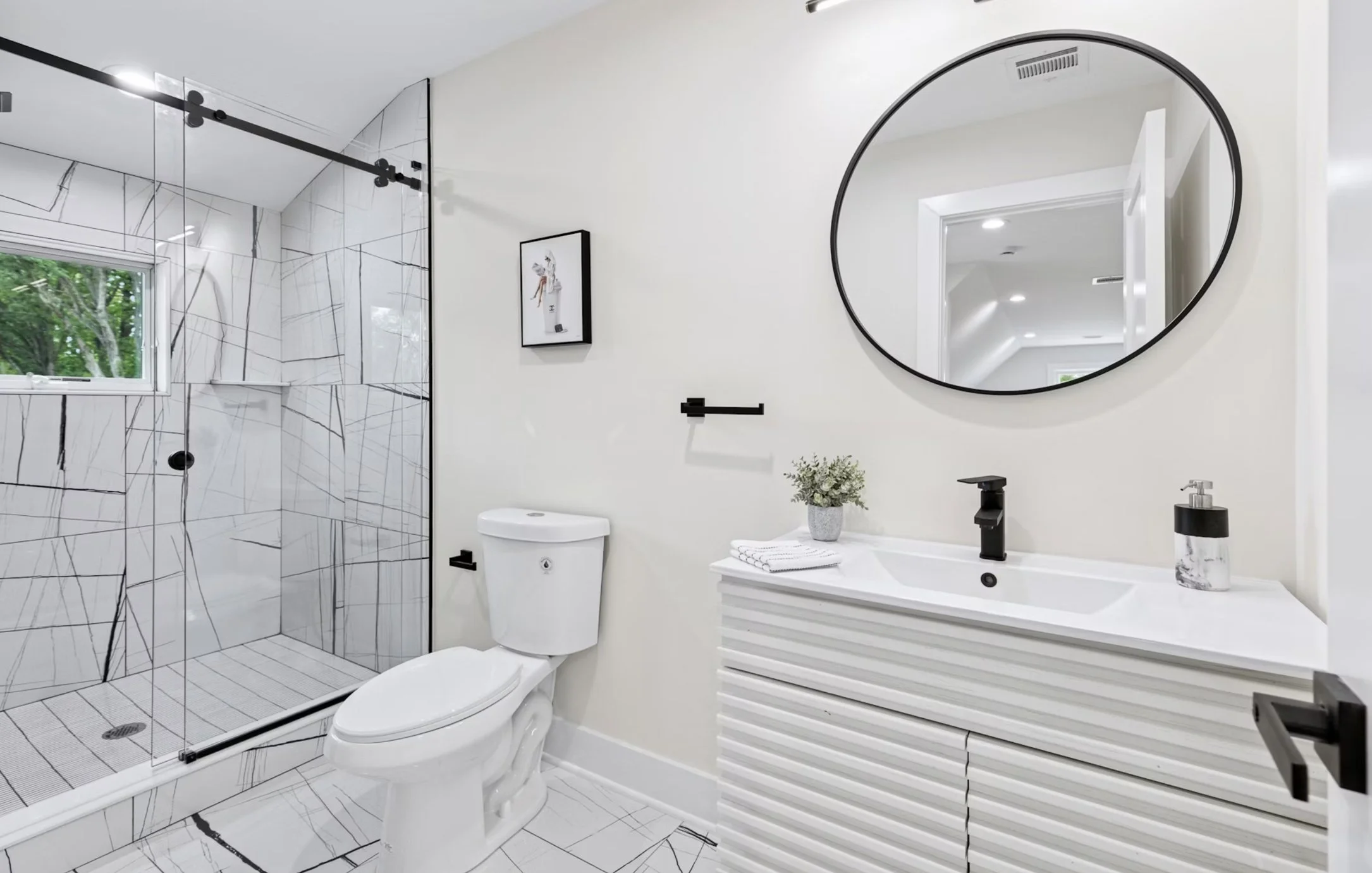 Modern bathroom with a walk-in shower, white ceramic toilet, and a white vanity with black fixtures. The shower has white tiles with black veining, a small window, and a glass door. A round mirror is above the vanity, and there is a small potted plan