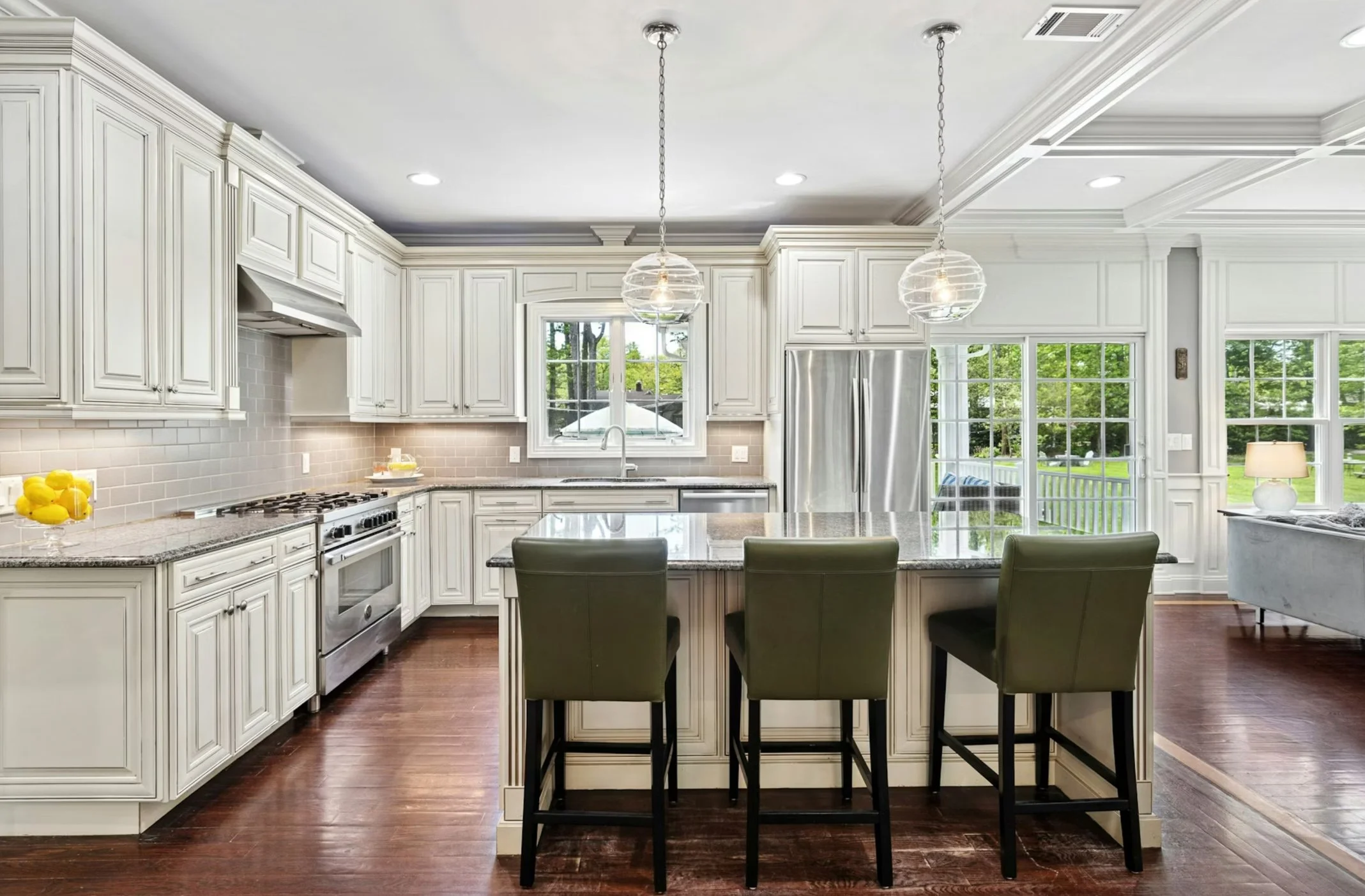 Modern kitchen with white cabinetry, stainless steel appliances, a large window, and a kitchen island with four green chairs. Hardwood floors and pendant lights hang above the island.