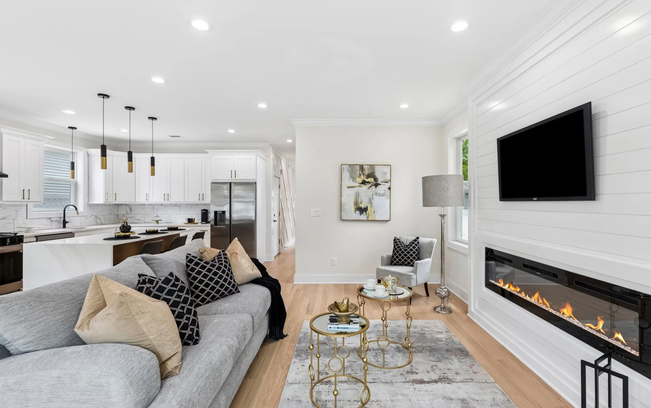 Modern open-concept living room with a gray sofa, black and beige pillows, gold and black side tables, a white stone fireplace, mounted TV, and dining area with a kitchen in background.