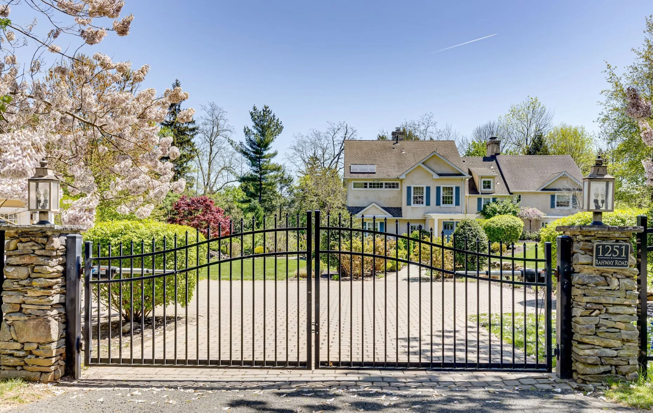 View of a large house behind a black wrought iron gate with stone pillars, surrounded by trees and a well-maintained garden, on a sunny day.