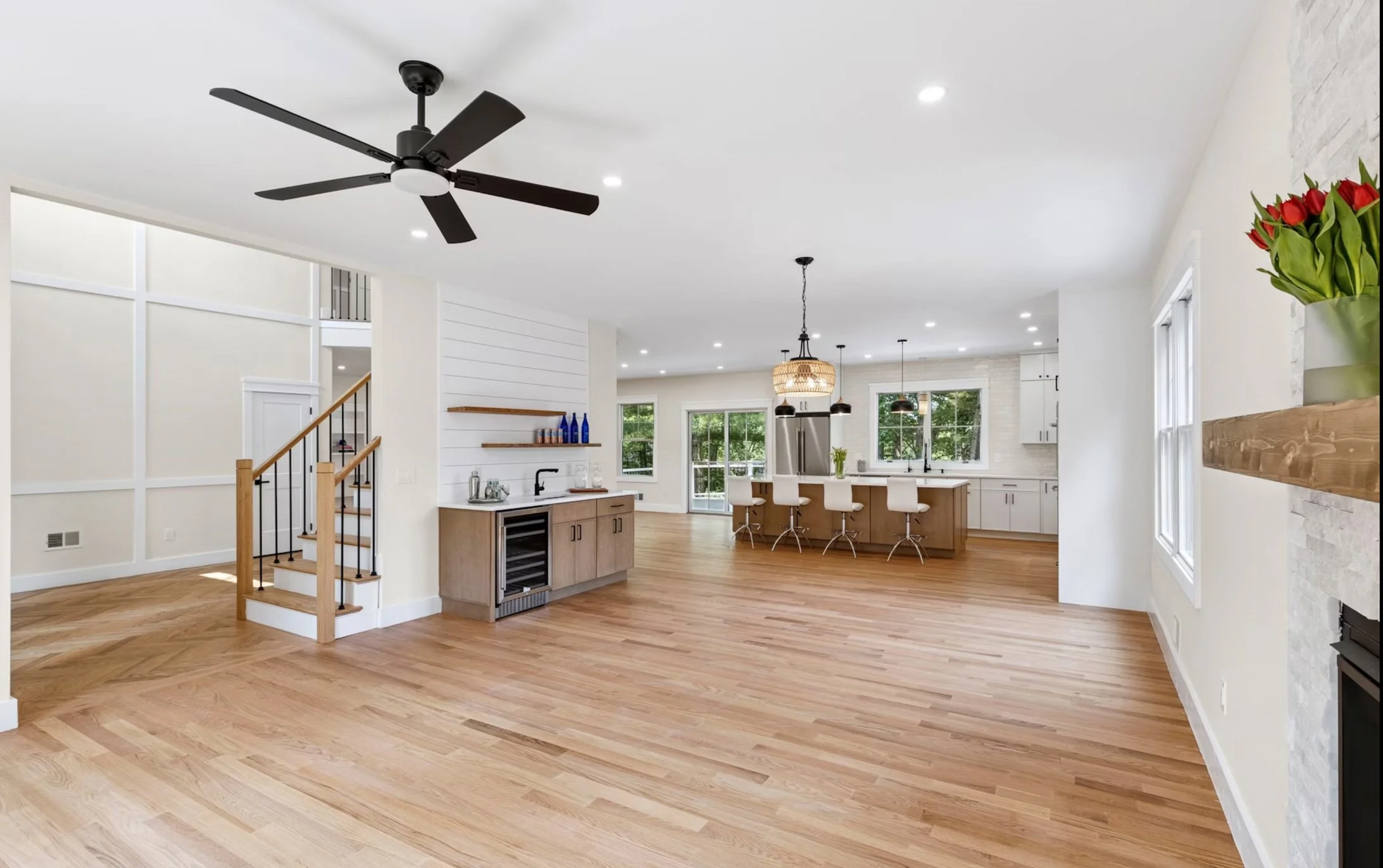 Open concept living area with hardwood floors, kitchen island with bar stools, white cabinets, large windows, and a fireplace with a wooden mantel and a vase of red tulips.