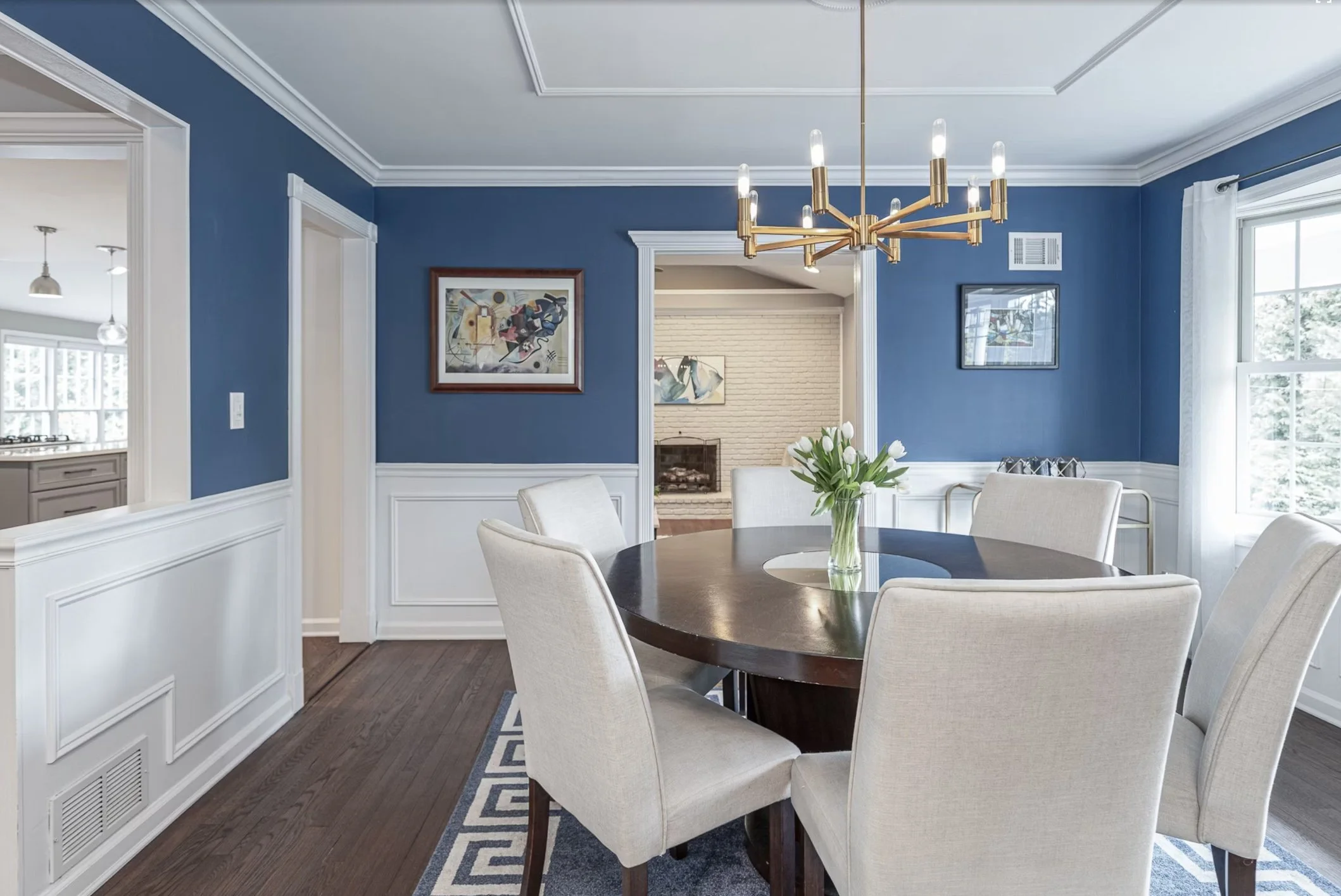 Dining room with blue walls, white wainscoting, a dark wood round table, six cream-colored chairs, a vase of white tulips, a modern gold chandelier, and large windows letting in natural light.