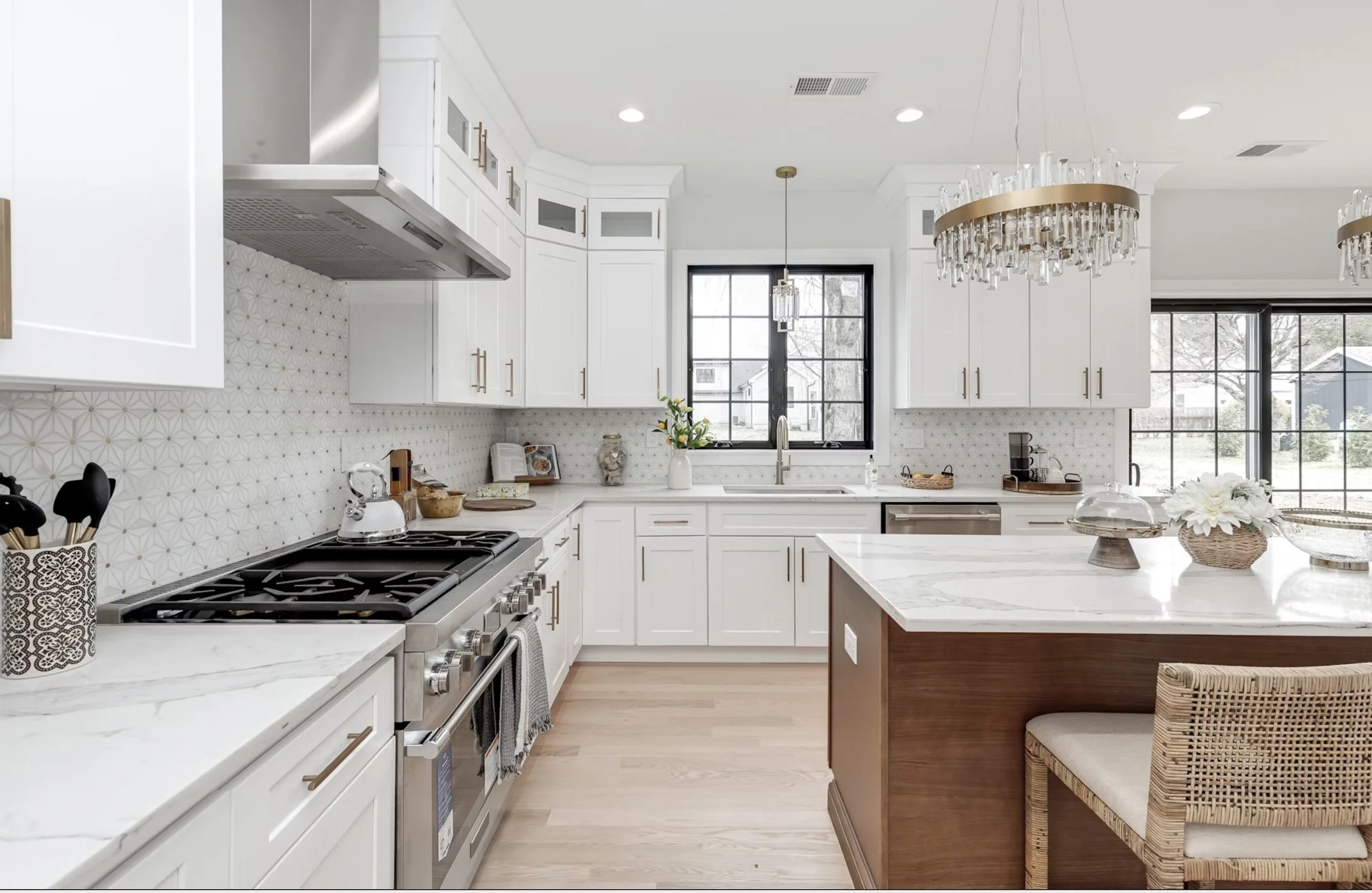 Modern white kitchen with marble countertops, black-framed window, hanging chandeliers, wooden island with a wicker chair, and decorative accents.