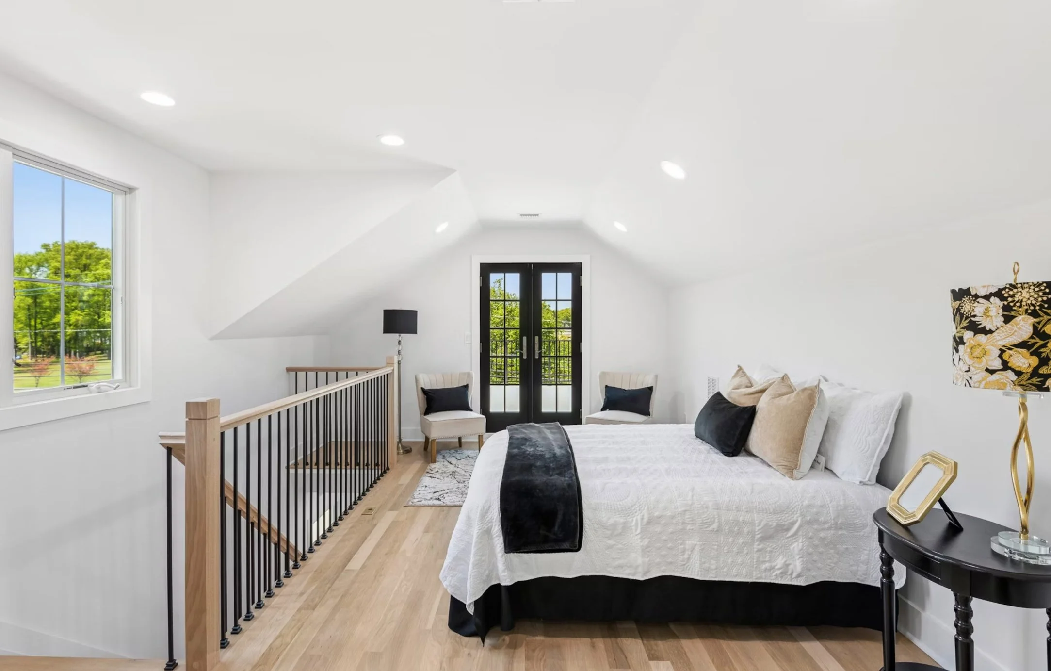 A bright, modern bedroom with white walls and ceiling, hardwood floors, and black and white furniture. A large bed with white bedding and black and beige pillows is in the center. A black nightstand with a decorative lamp and picture frame is beside 