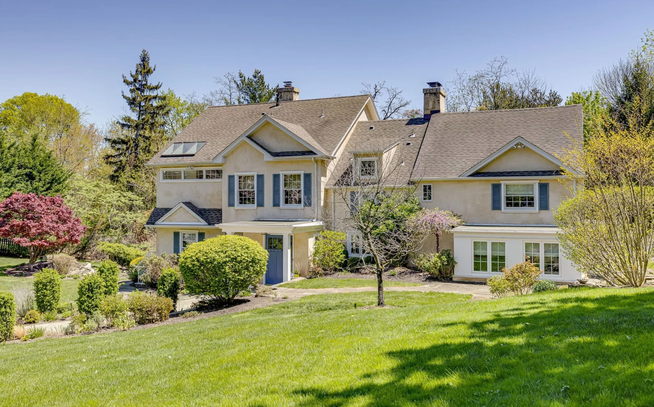 A large, multi-story house with a well-maintained front yard, lush green grass, various trees, and shrubs, with a clear blue sky in the background.