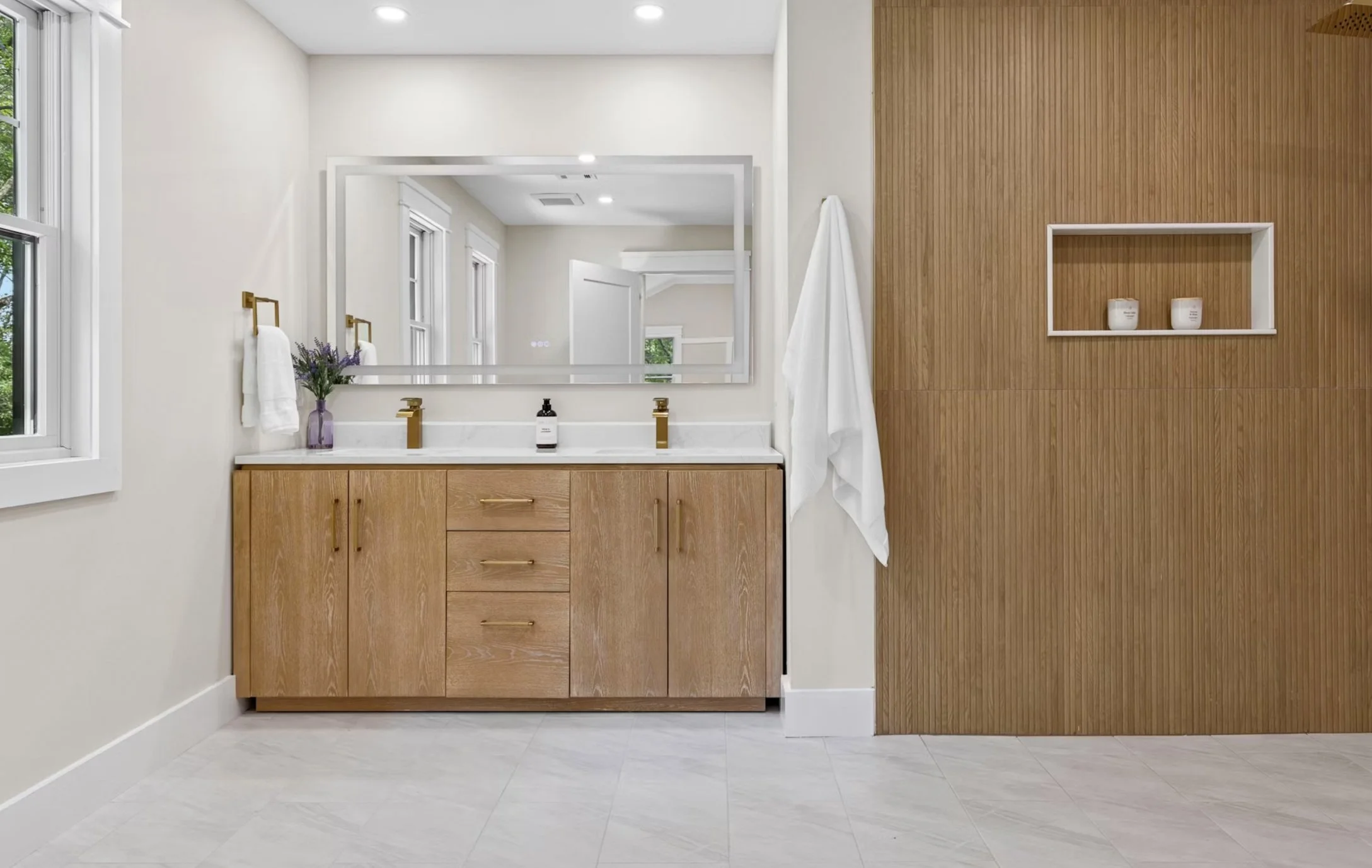 Modern bathroom with wooden vanity, large mirror, white walls, and a wooden accent wall with built-in shelving containing small candles.