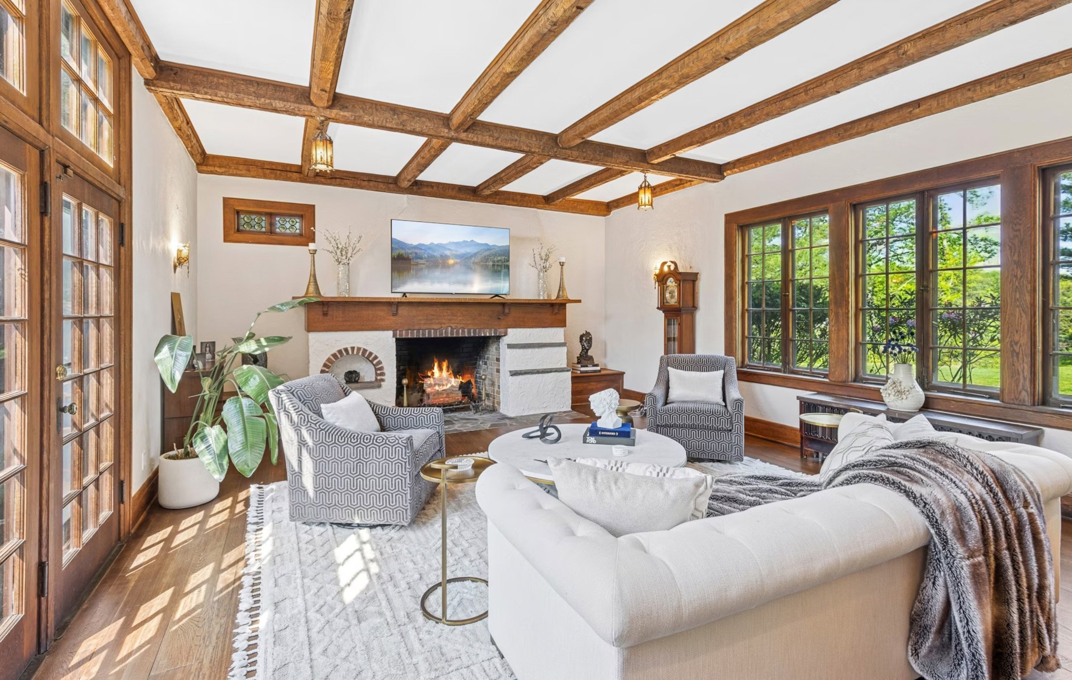 Living room with white walls, wooden ceiling beams, large windows, a fireplace, and contemporary furniture including a white sofa, patterned armchairs, and a coffee table with decor items.