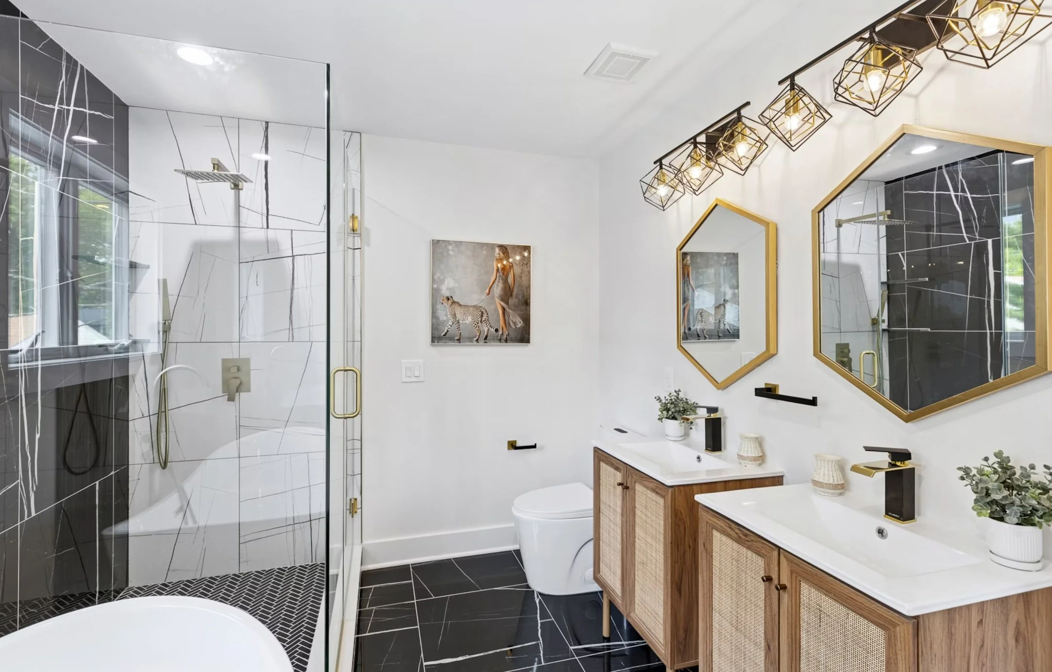 Modern bathroom with black and white marble tiles, double vanity with white sinks and wooden cabinets, gold-framed mirrors, black and gold fixtures, a glass shower with black and white marble tiles, and two framed pictures on the wall.