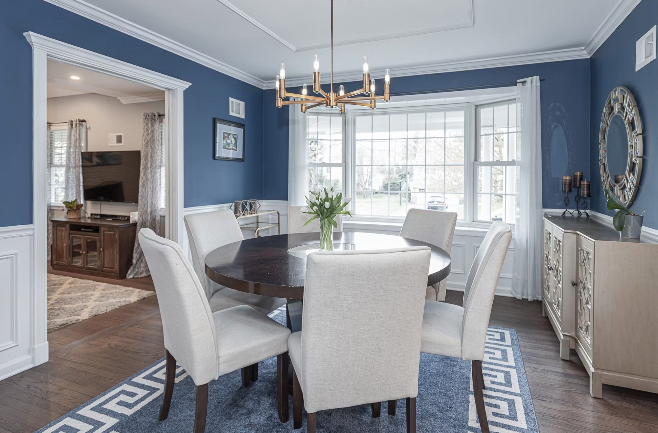 Dining room with blue walls, white wainscoting, and a round dark wood table with six white upholstered chairs. A gold chandelier hangs above, and large windows with sheer white curtains let in natural light. There is a white sideboard with decorative