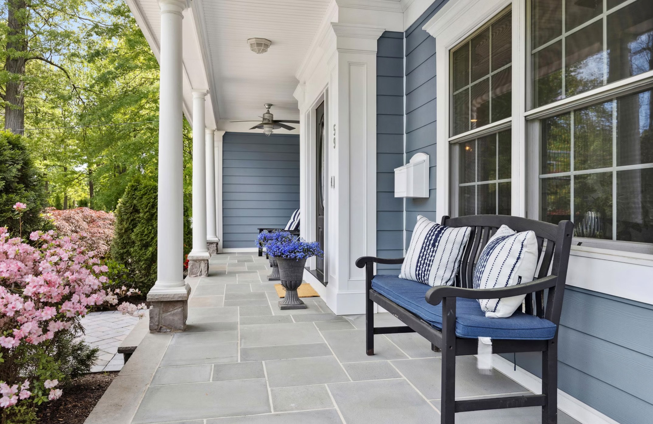 Front porch of a house with blue siding, white trim, and large windows, decorated with a black bench with pillows, potted plants, and surrounded by flowering bushes.