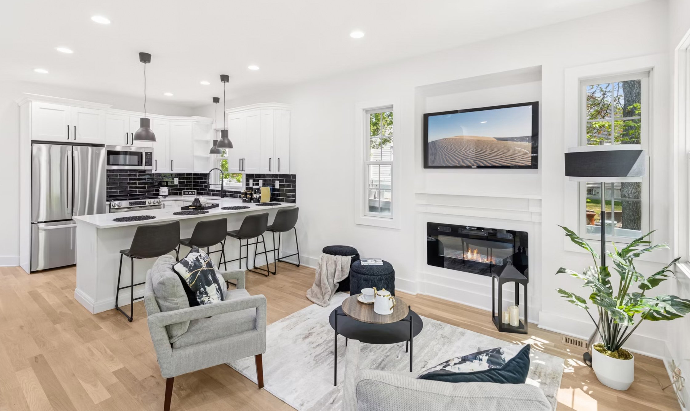 Open-concept living room and kitchen with white walls, hardwood floors, gray furniture, black accents, fireplace, wall-mounted TV, large windows, and houseplants.