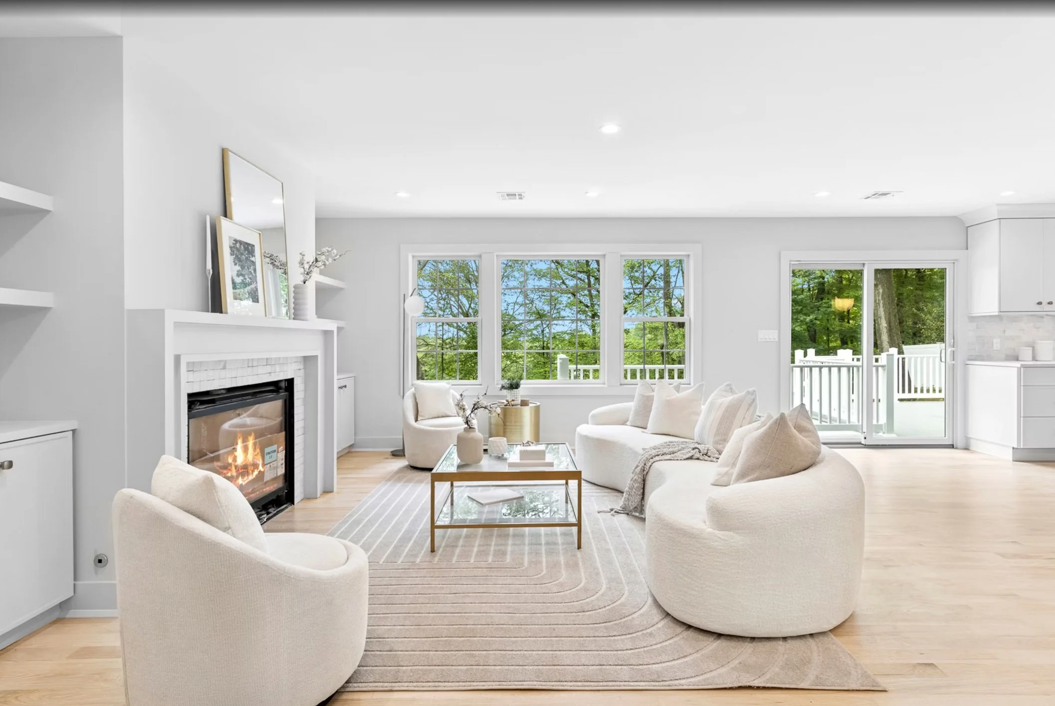 Bright living room with white walls, large windows showing green trees outside, a fireplace with a fire, and white furniture including a curved sofa and armchair, with a glass coffee table on a beige striped rug.