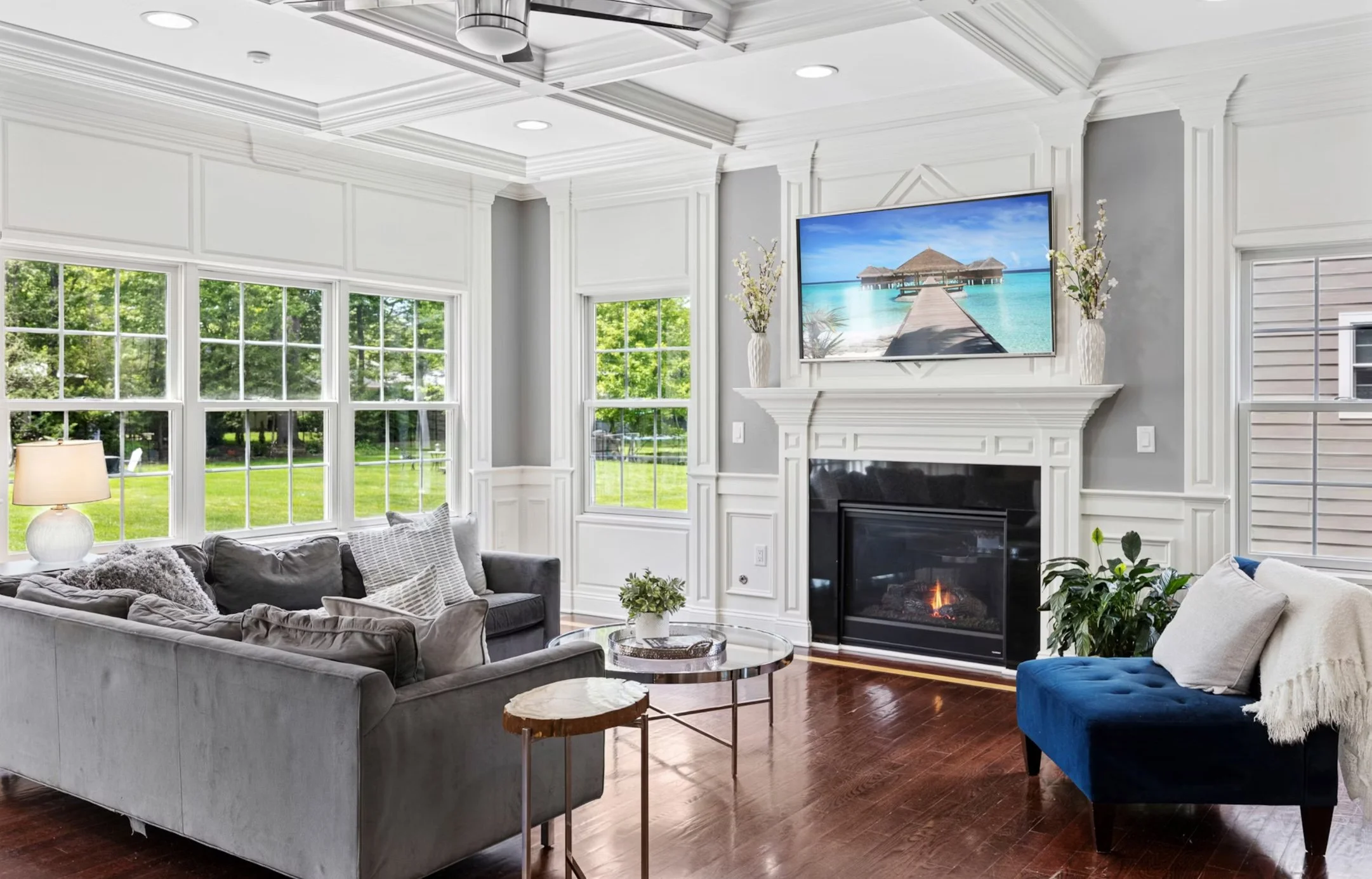 Living room with large windows, white walls and trim, a fireplace, a wall-mounted TV, a gray sofa with pillows, a blue bench with a throw blanket, a round side table, a small plant, and a landscape view outside.
