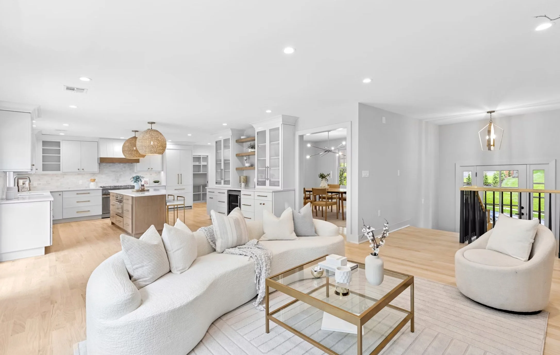 Open-concept living room and kitchen with modern decor, white walls, light wood flooring, white sofa with pillows, circular armchair, glass-top coffee table, wooden dining table in the background, and large windows bringing in natural light.