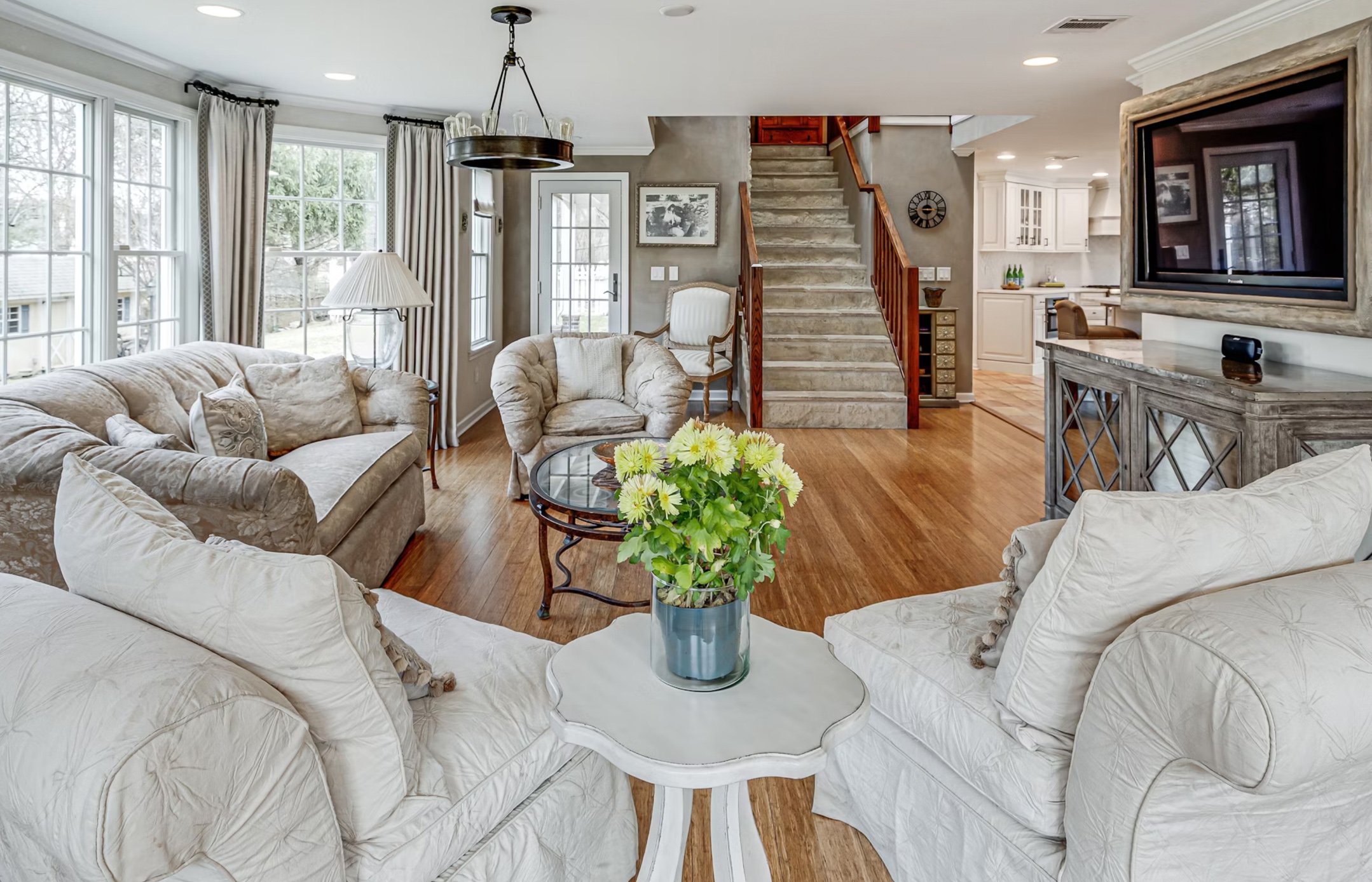 Living room with beige sofas and chairs, wooden floors, large windows with curtains, a round coffee table with a vase of yellow flowers, a staircase with wooden railing, and a TV on a console.