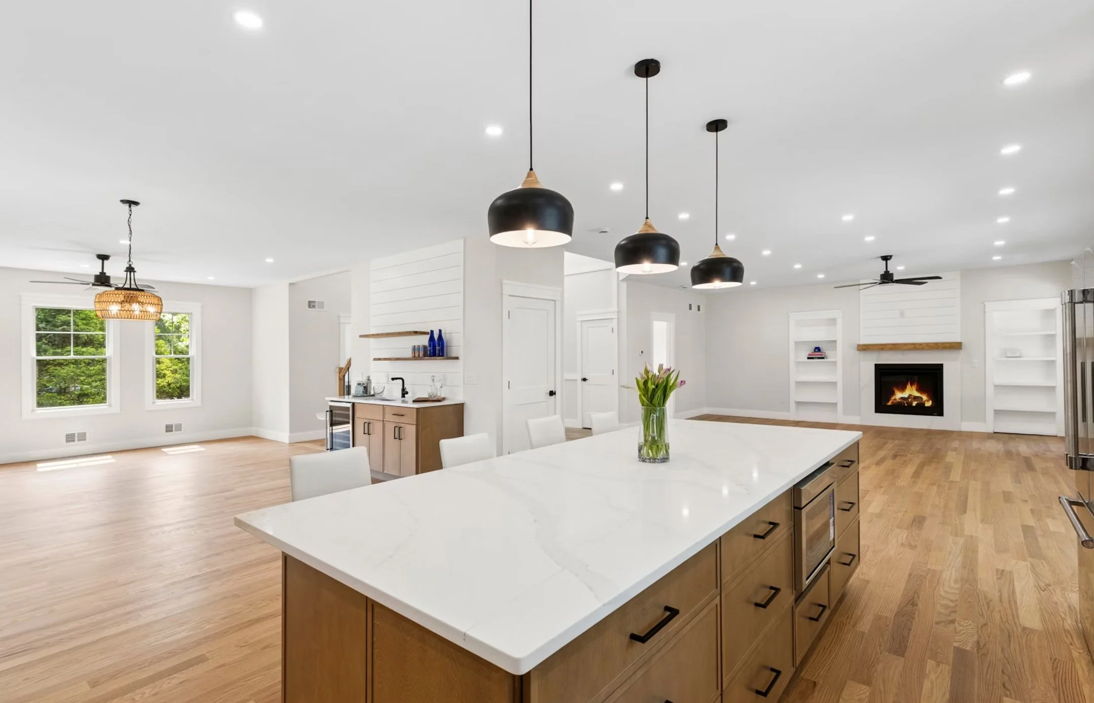 Open-concept living space with a kitchen island, white walls, hardwood floors, large windows, and a fireplace with built-in shelves.
