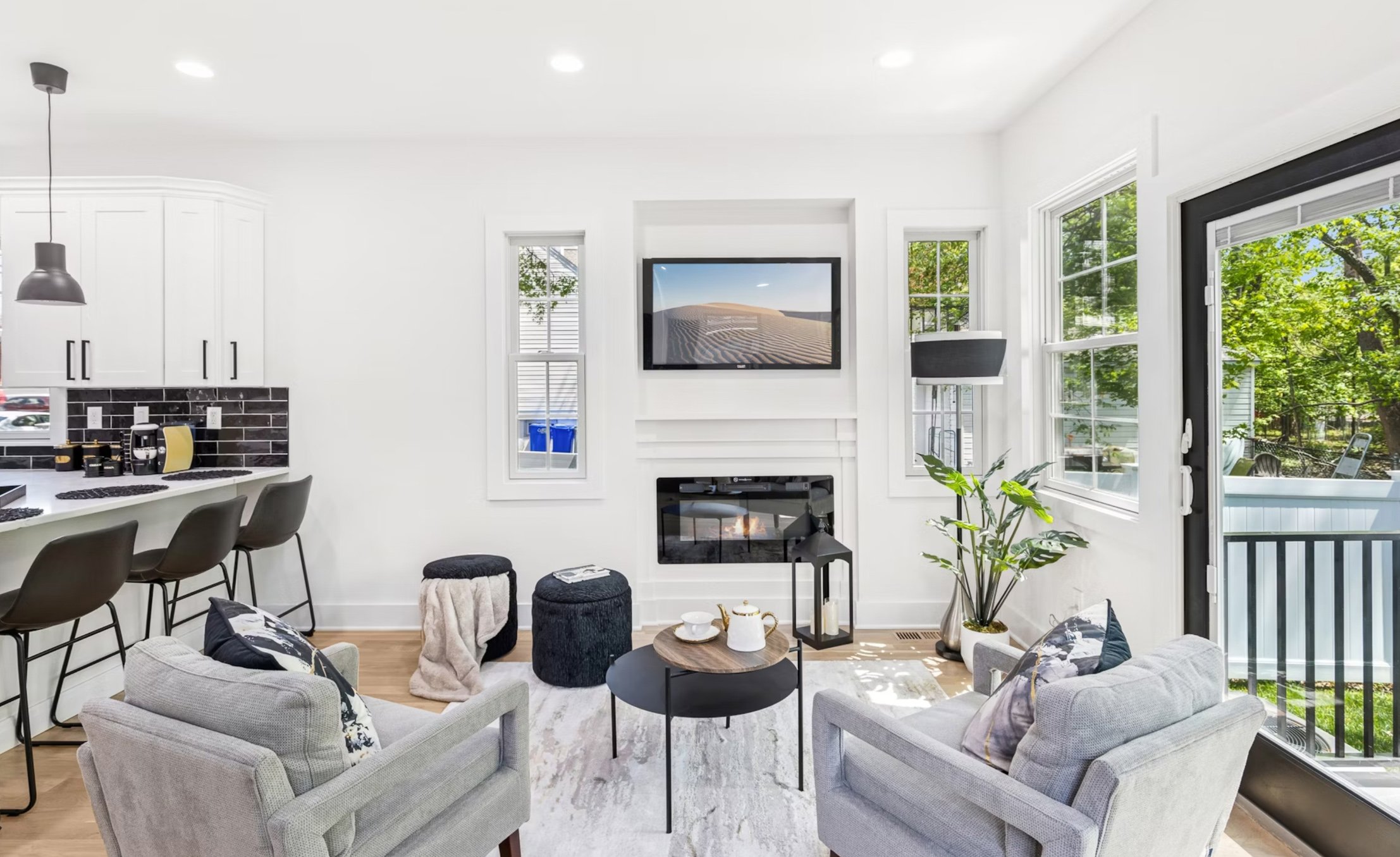 Bright living room with two gray armchairs, a black coffee table, a fireplace, a wall-mounted TV, large windows, and a sliding glass door leading outside.