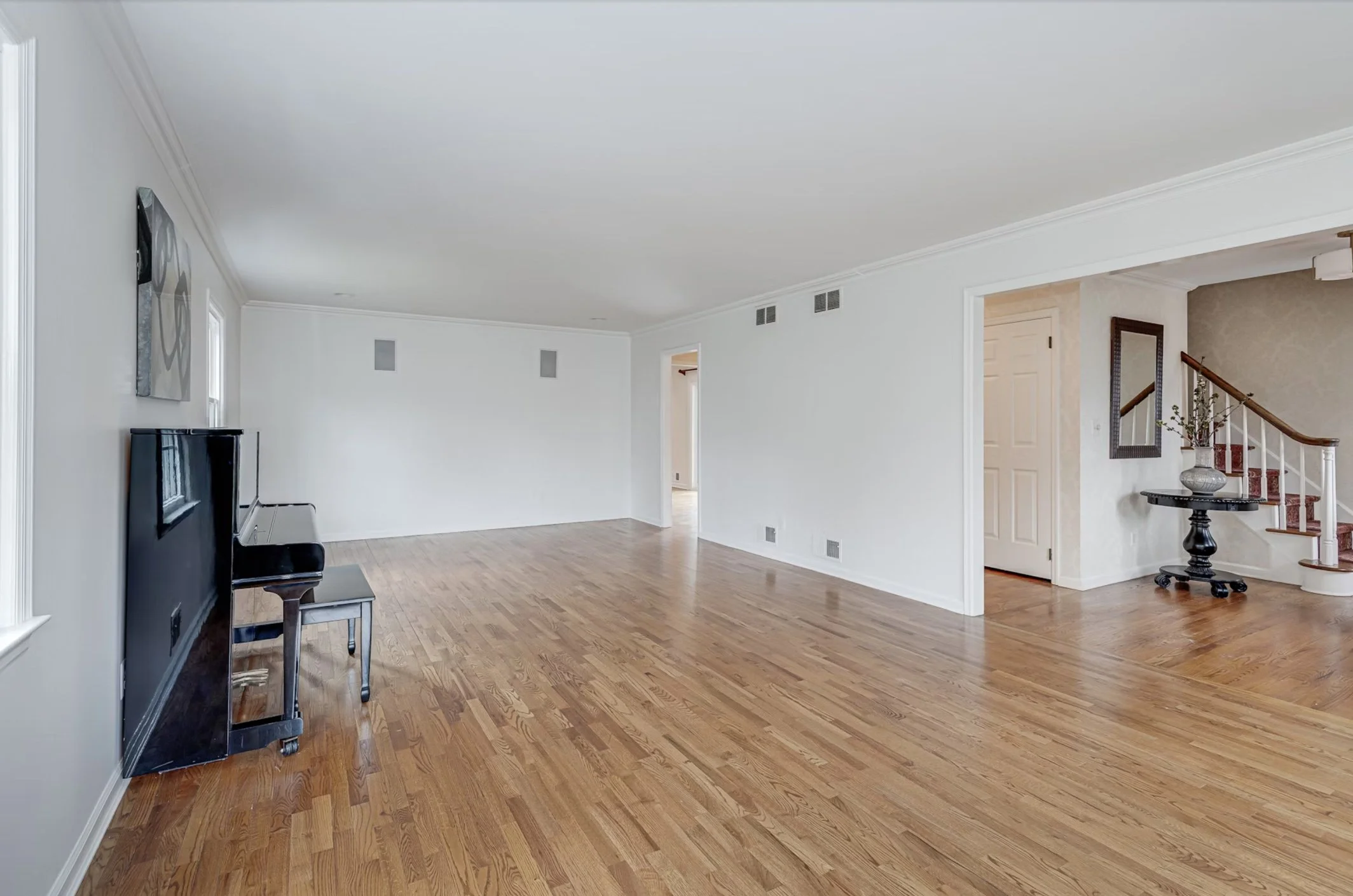 Empty living room with hardwood floors, white walls, and a staircase with a wooden handrail and red carpeted stairs, decorated with a vase of flowers. A piano with a bench is visible near the windows.