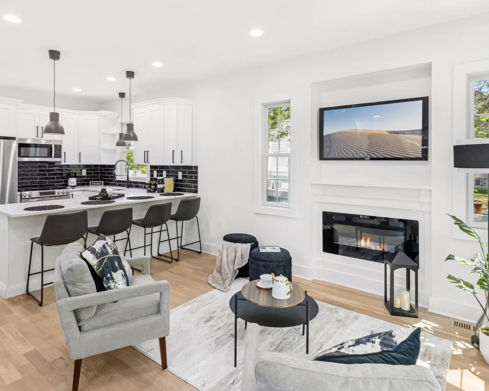 Modern open-concept living room and kitchen with white walls, black and white decor, and wooden flooring. The living room features a wall-mounted TV above a fireplace, a light-colored armchair with a decorative pillow, and a coffee table with a teapot and cups. The kitchen has white cabinets, black backsplash tiles, a stainless steel refrigerator, microwave, and stove, and a breakfast bar with four black chairs.