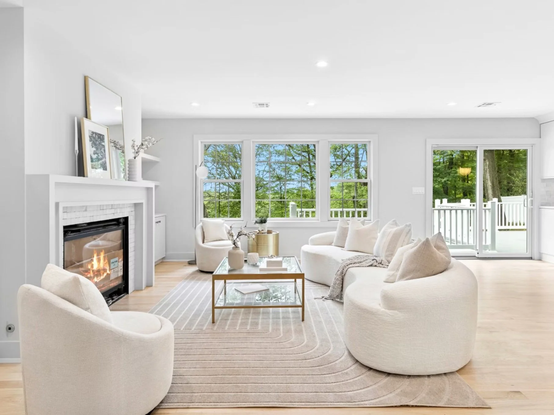 Bright living room with white walls, light wood flooring, a white sofa set, a fireplace with a fire, a glass coffee table, and large windows showing green trees outside.