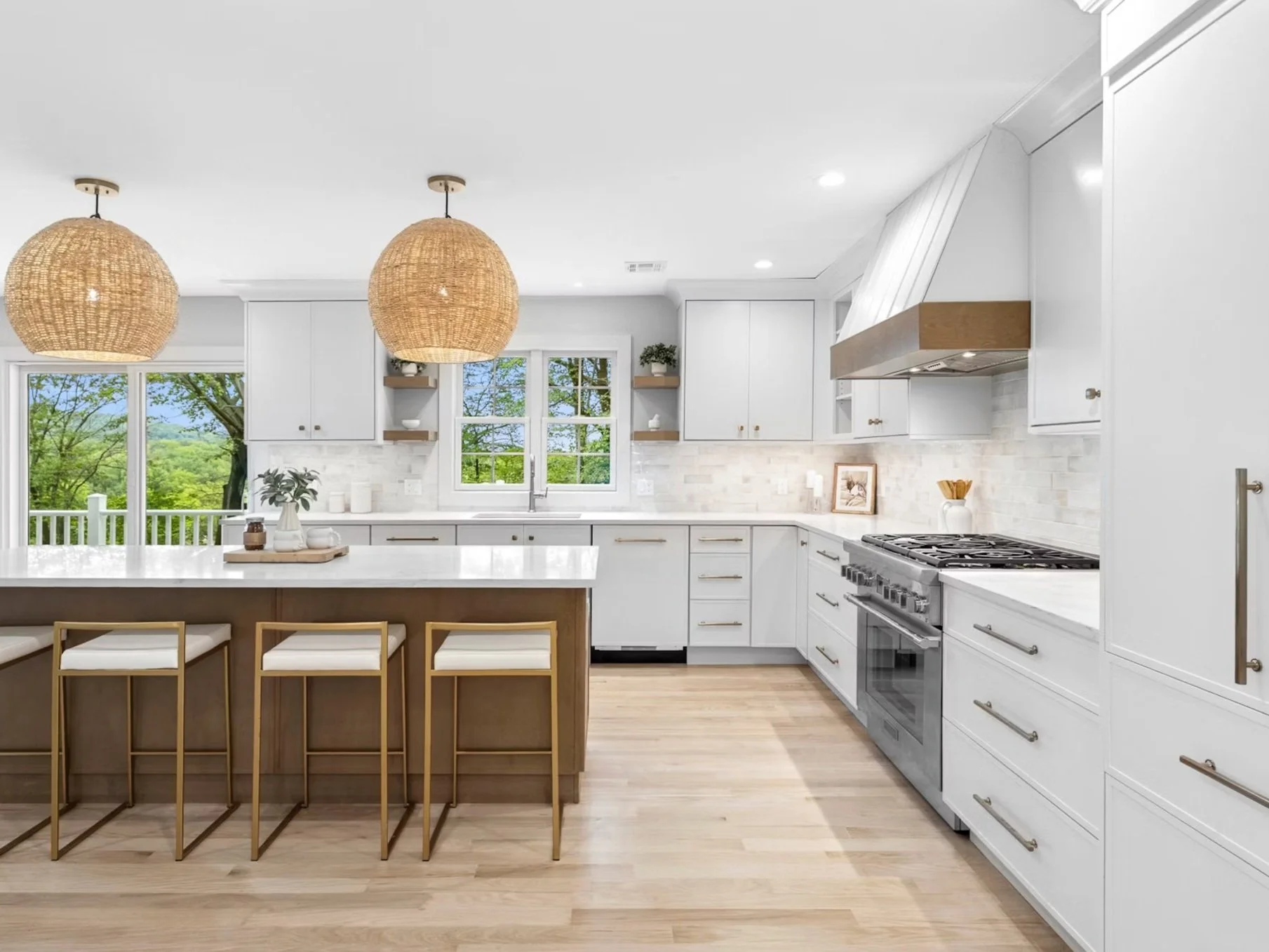 Modern kitchen with white cabinets, a large island with a white countertop, three gold bar stools, two woven pendant lights, wooden shelves, a gas stove, and a window overlooking green trees.