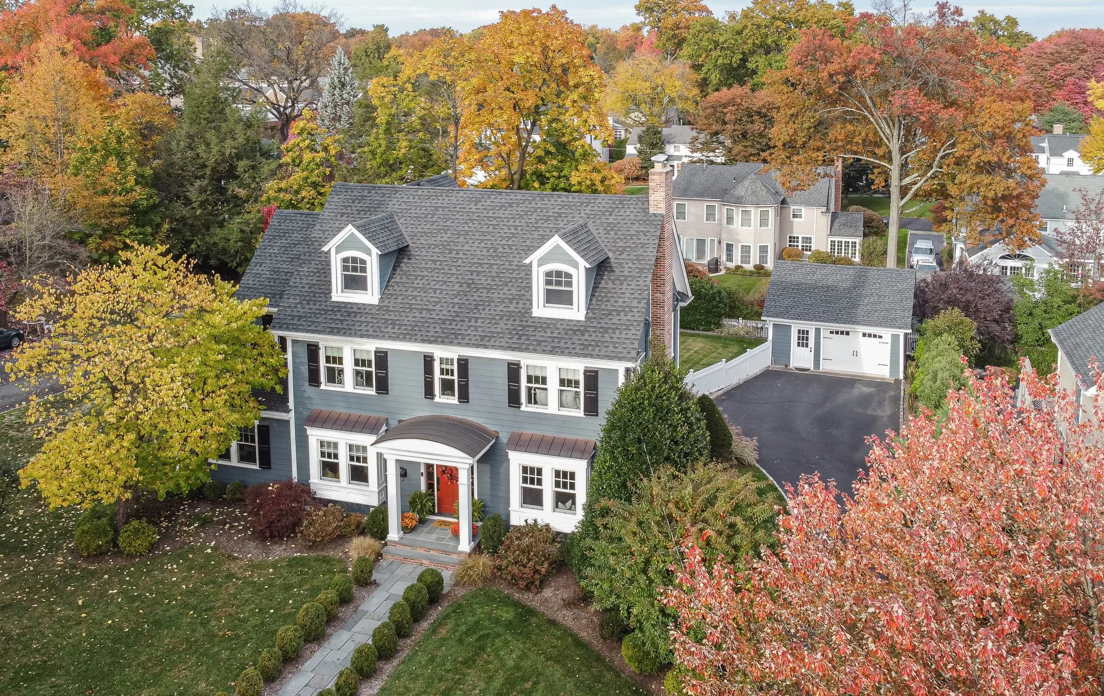 An aerial view of a two-story house with a gray roof and light blue exterior, surrounded by colorful fall trees, with a driveway and a detached garage in the backyard.