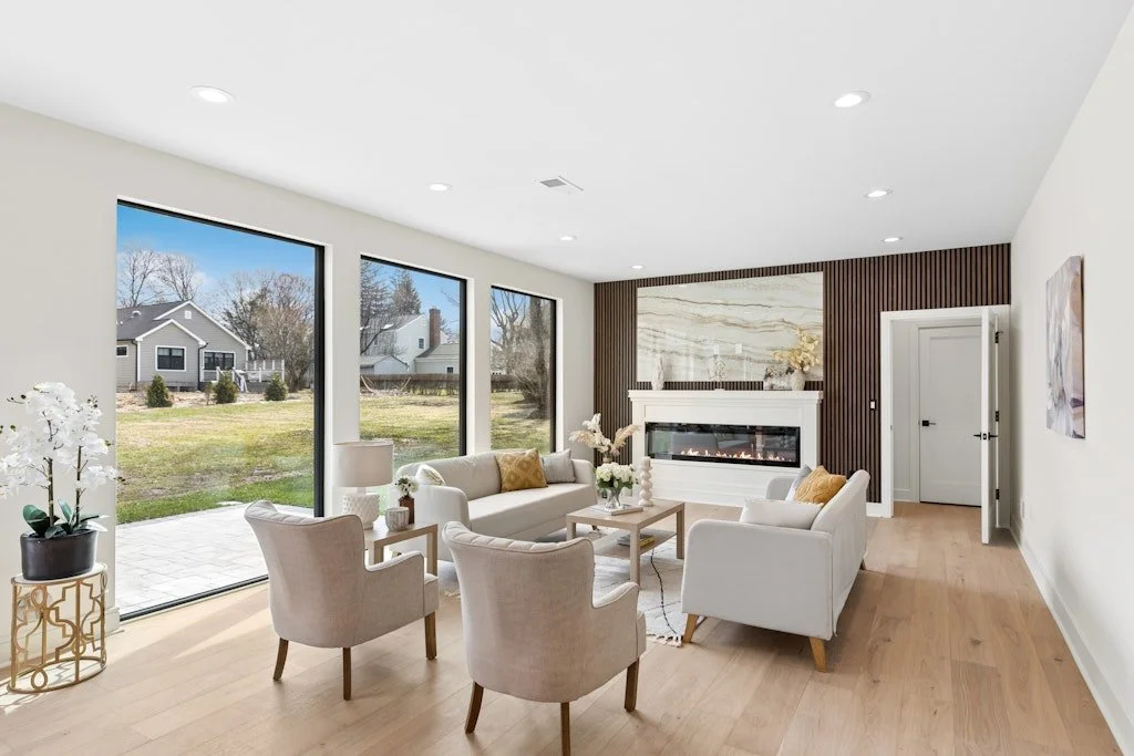 Living room with large windows, beige chairs and sofa, wooden floor, white walls, fireplace, and minimal decor.
