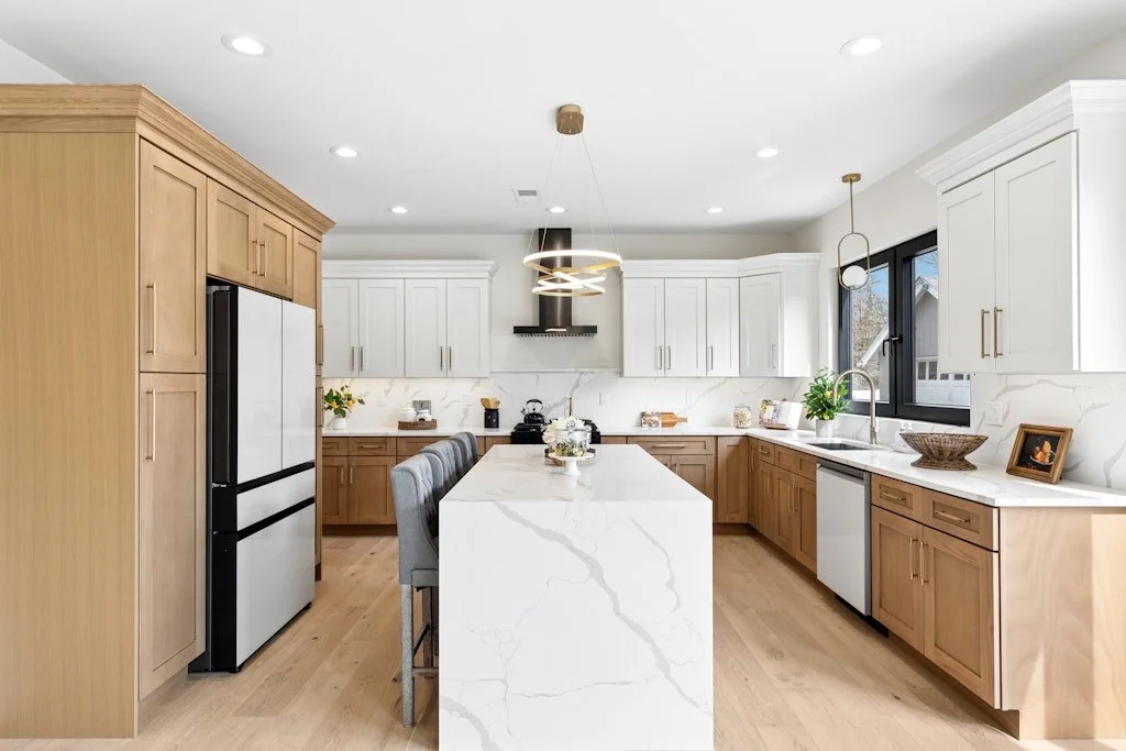 Modern kitchen with white and light wood cabinets, marble countertops, a large central island, stainless steel appliances, and natural light from a window.