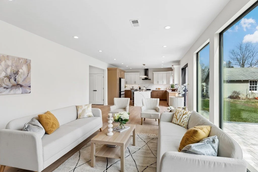 Bright open living room with white couches, pillows, and a coffee table, adjacent to a kitchen with white cabinets and wooden accents, large sliding glass doors revealing a backyard with grass and trees.
